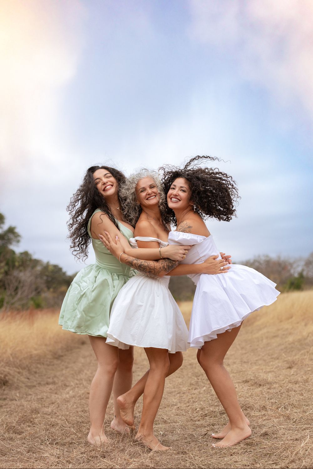 Three friends in white and mint green dresses laugh and embrace while standing barefoot on a grassy hillside.