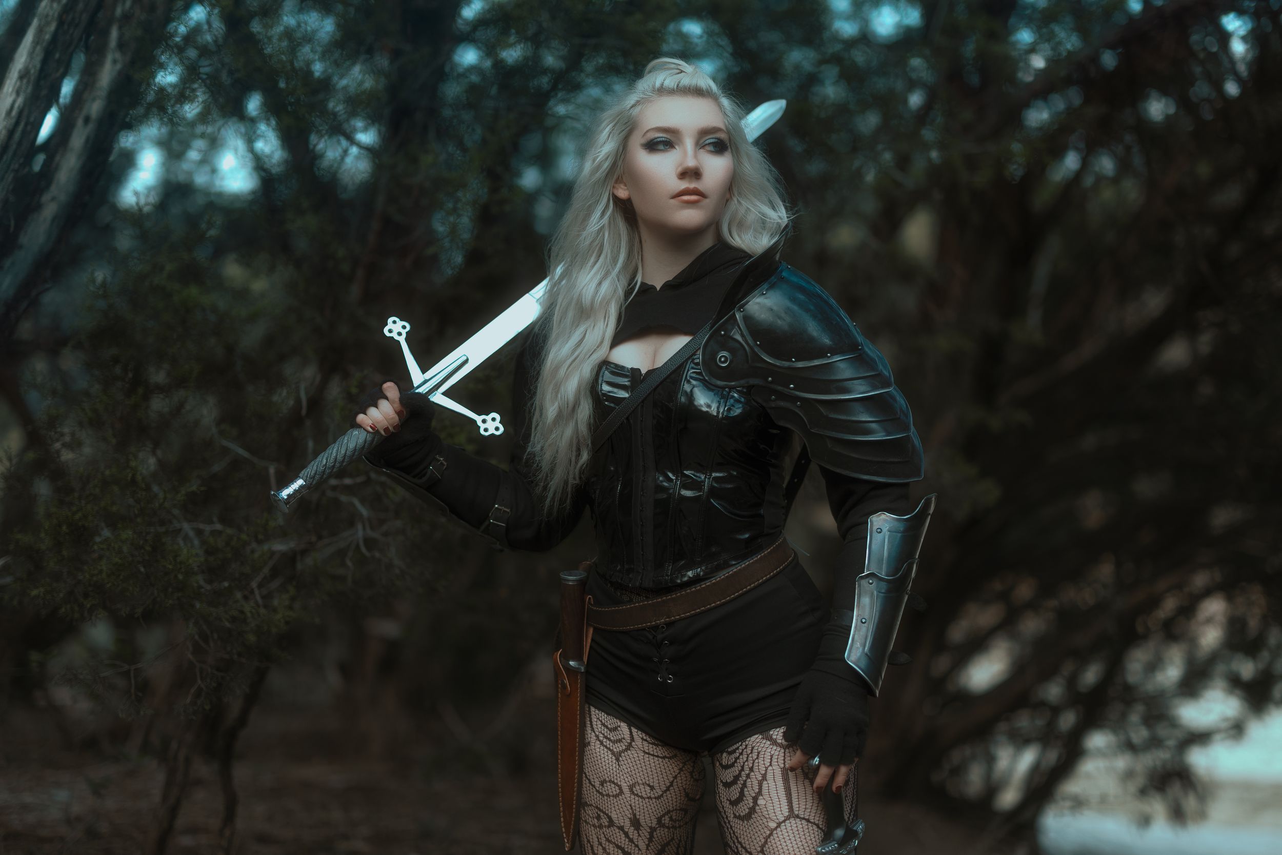 Woman in dark elf cosplay with white hair, pointed ears, armor, and sword posing in a forest setting.