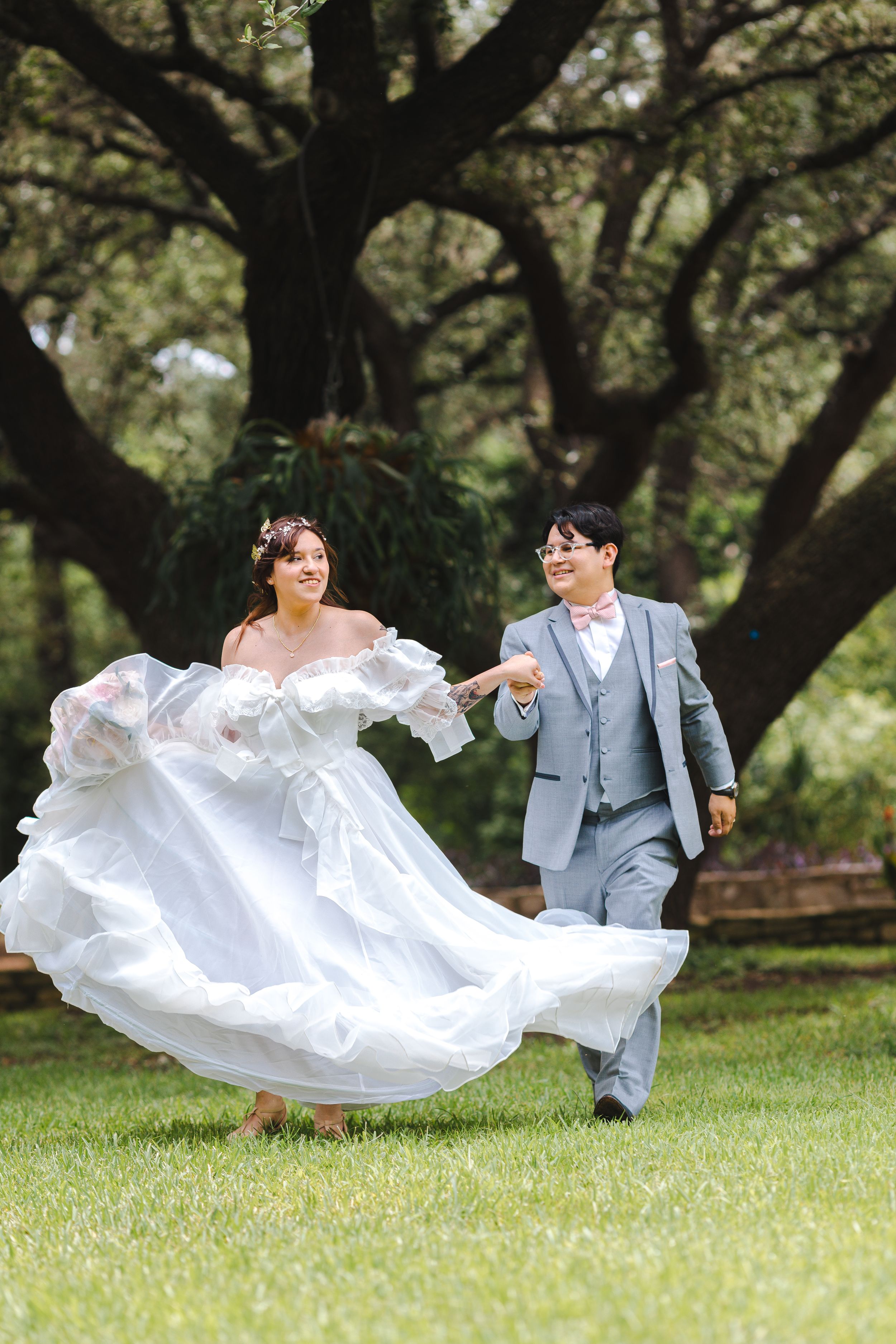 A couple in wedding attire playfully levitates on lush green grass beneath sprawling tree branches.