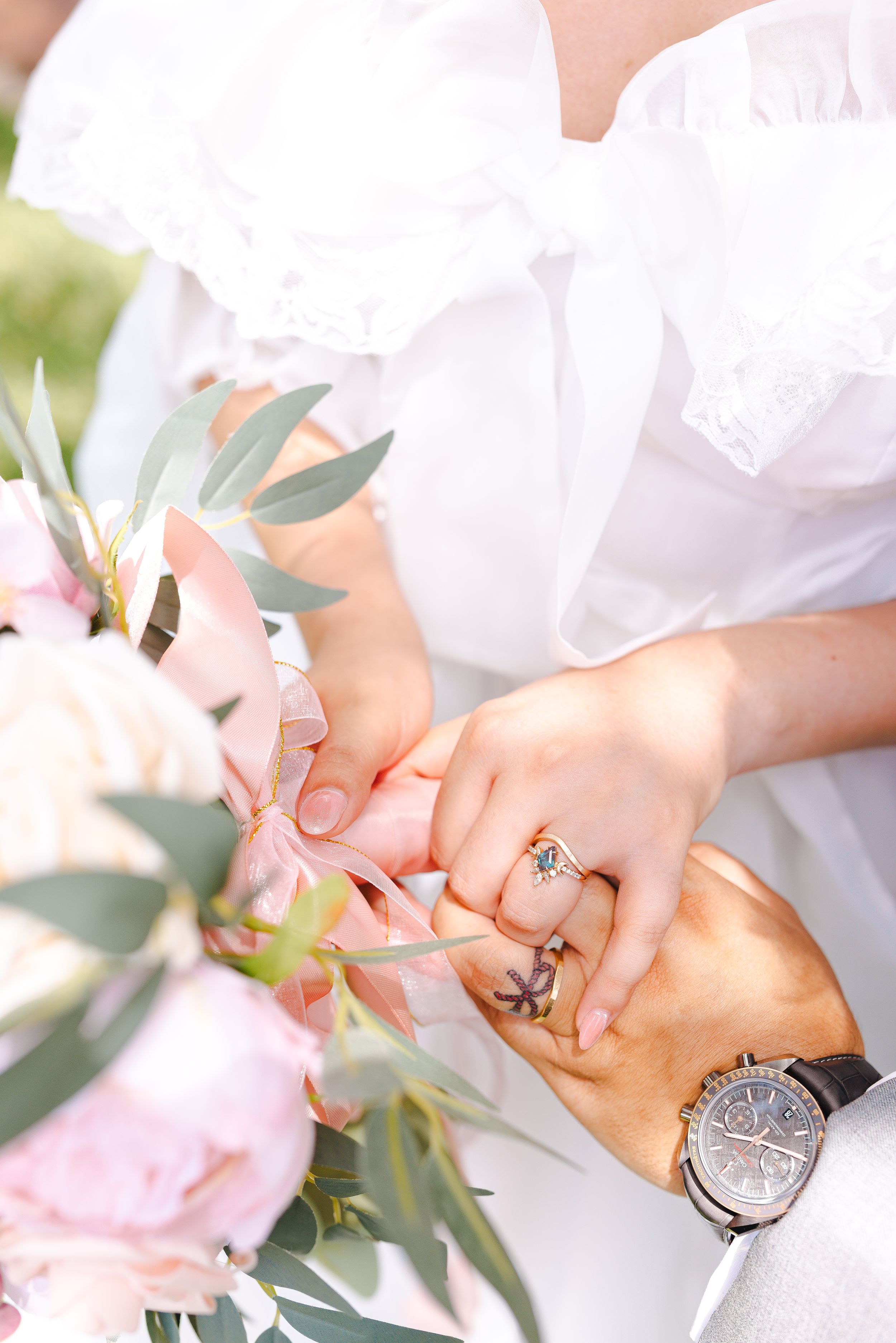 Close-up view of delicate hands with engagement ring and watch against soft pink floral background.