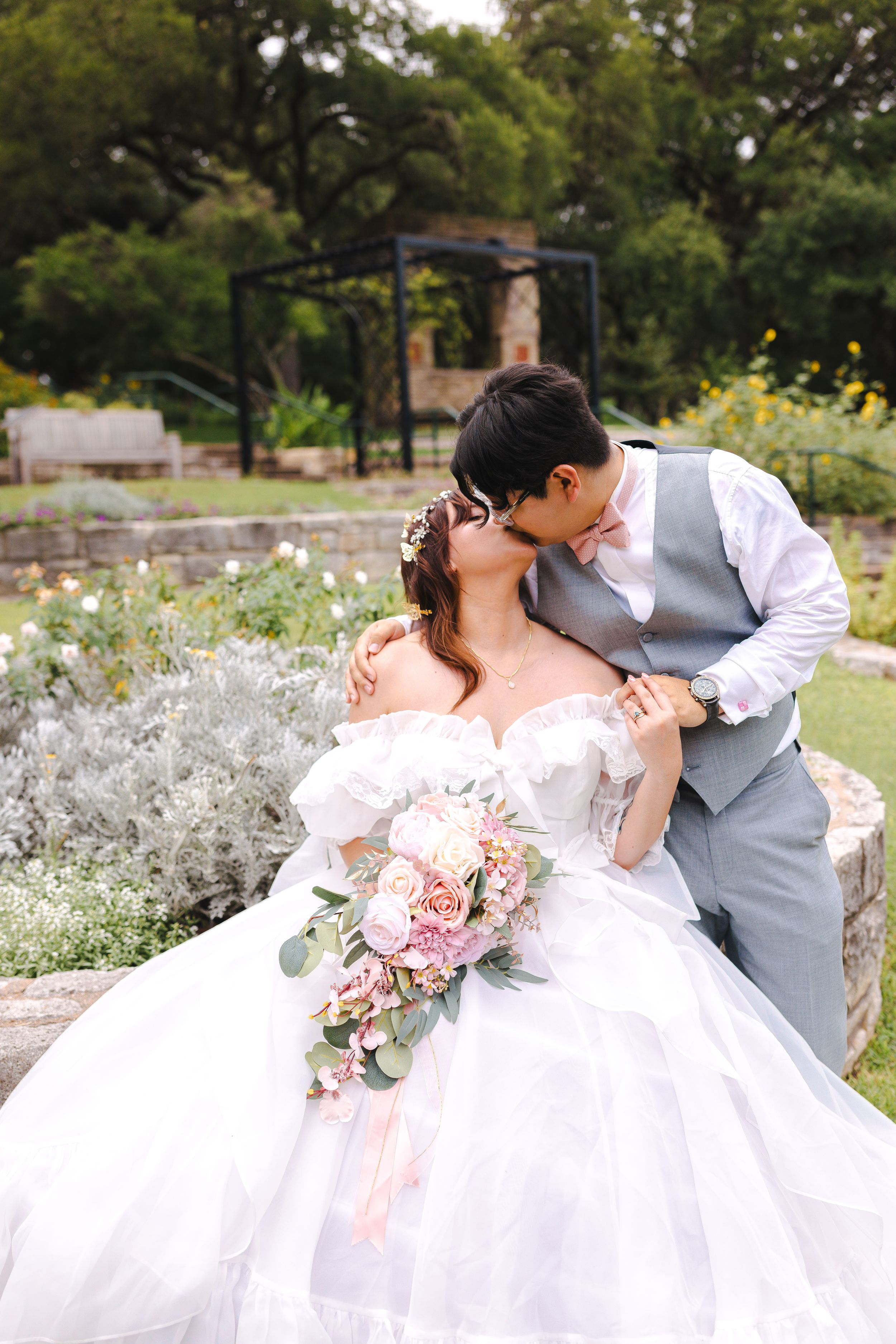 A tender wedding moment captured in a garden with white and pink flowers and a decorative stone wall.