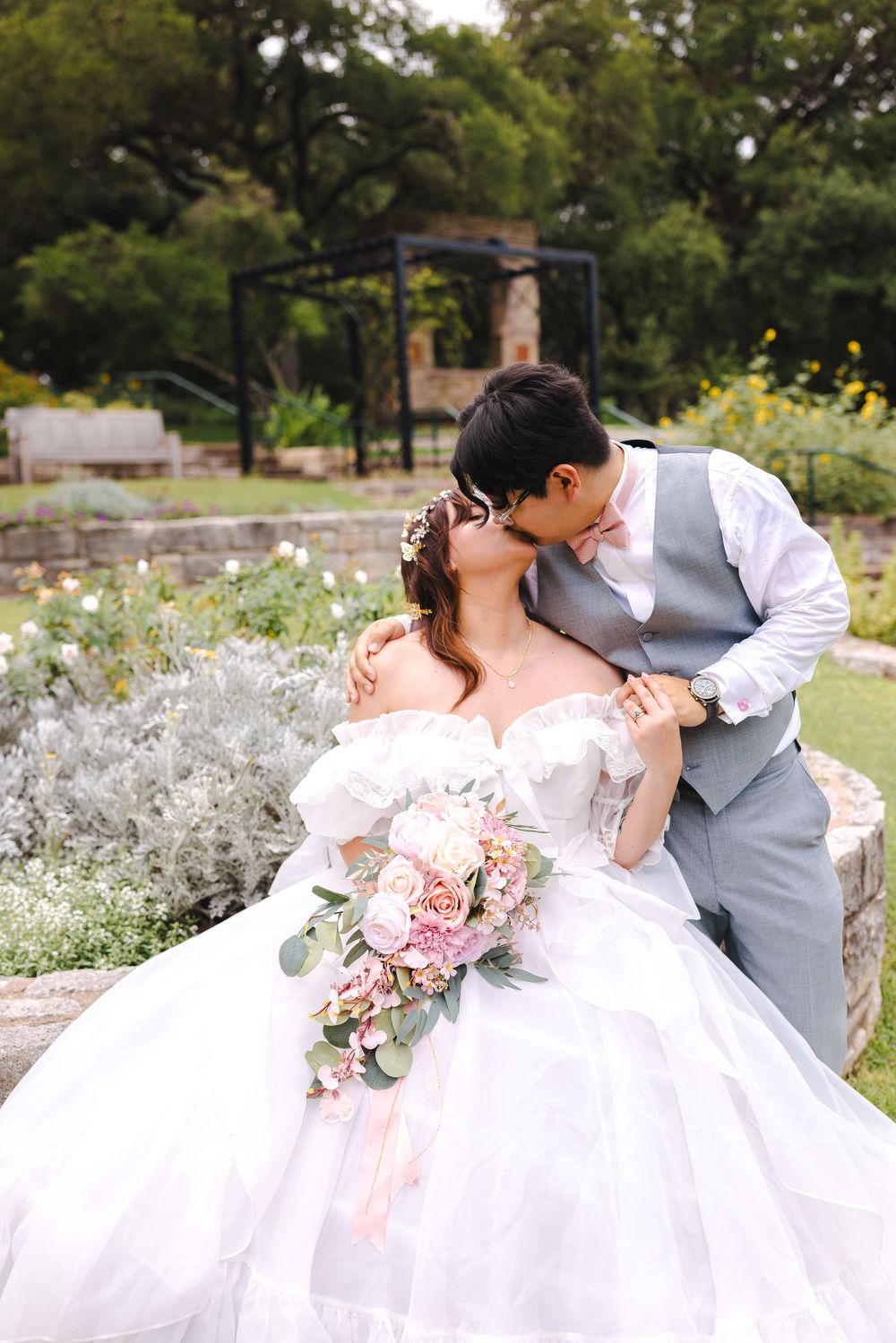 A tender wedding moment captured in a garden with white and pink flowers and a decorative stone wall.