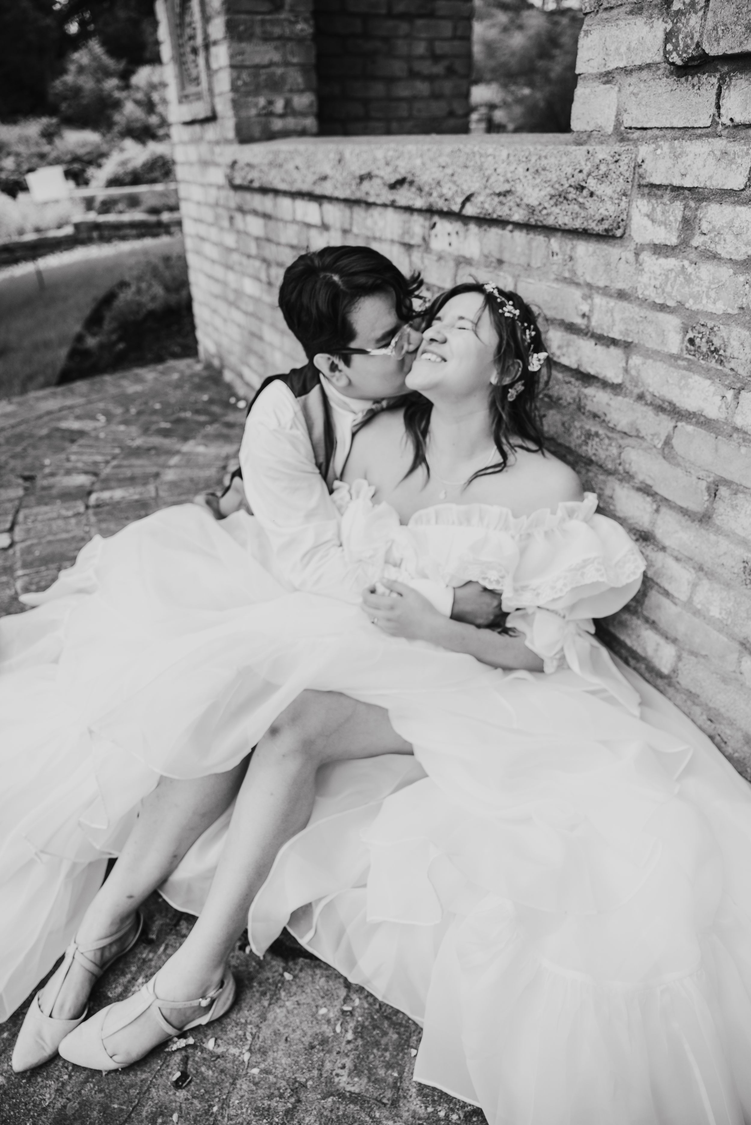 A romantic black and white photo series of a couple in wedding attire reclining against a brick wall.