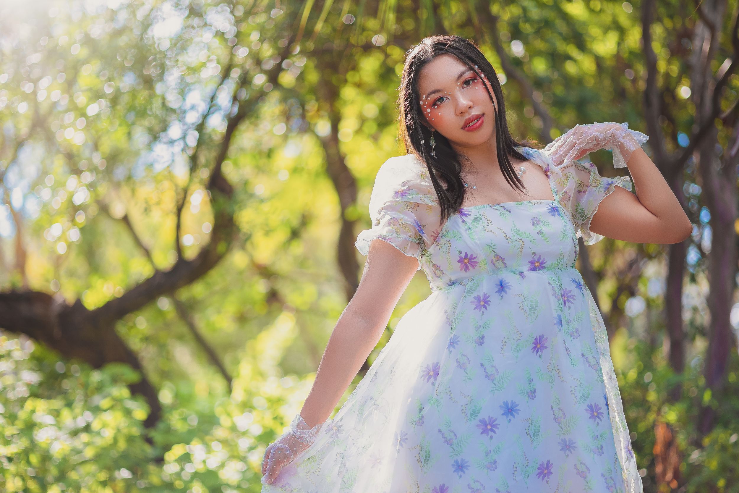 Young woman in floral white dress posing gracefully outdoors among sunlit trees in a garden setting.