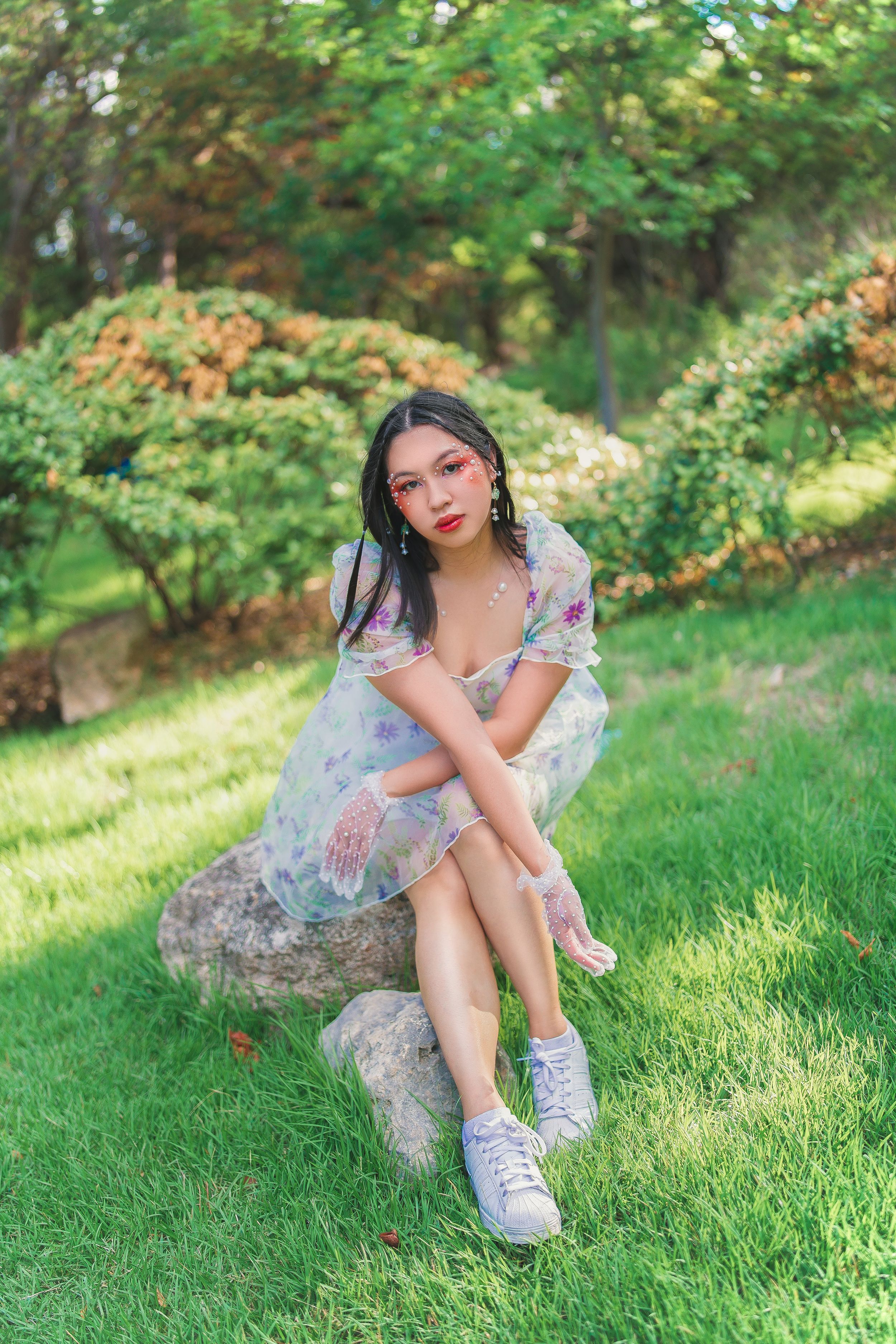 Young Asian woman in floral dress and white sneakers sitting on a rock in a lush green garden park.
