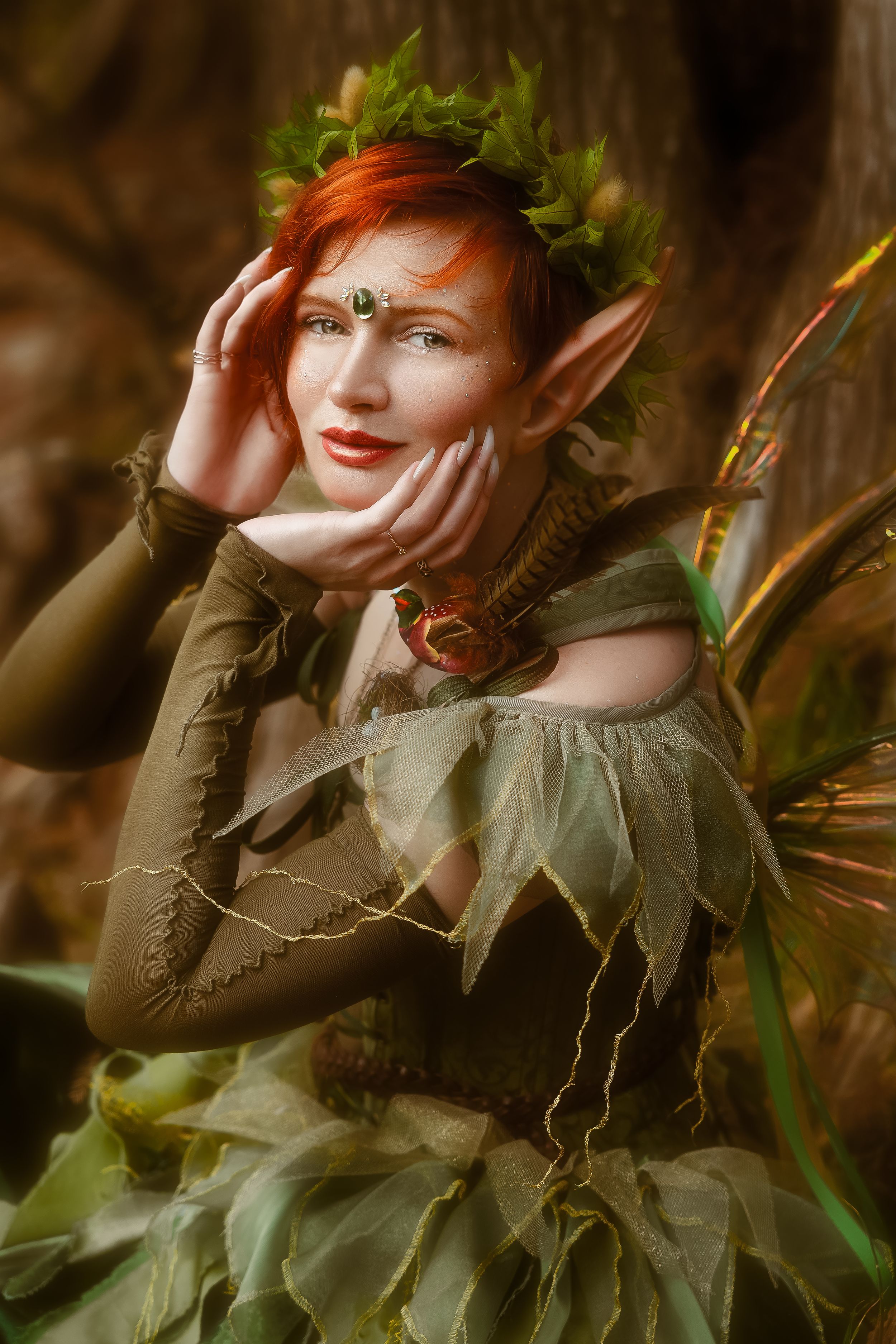 A red-haired woman in a green fairy costume with pointed ears, leaf crown, and wings poses in a forest setting.