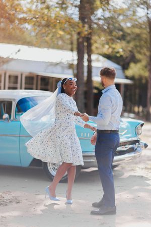 A couple in wedding attire dance together in front of a vintage blue Chevrolet car on a sunny autumn day.