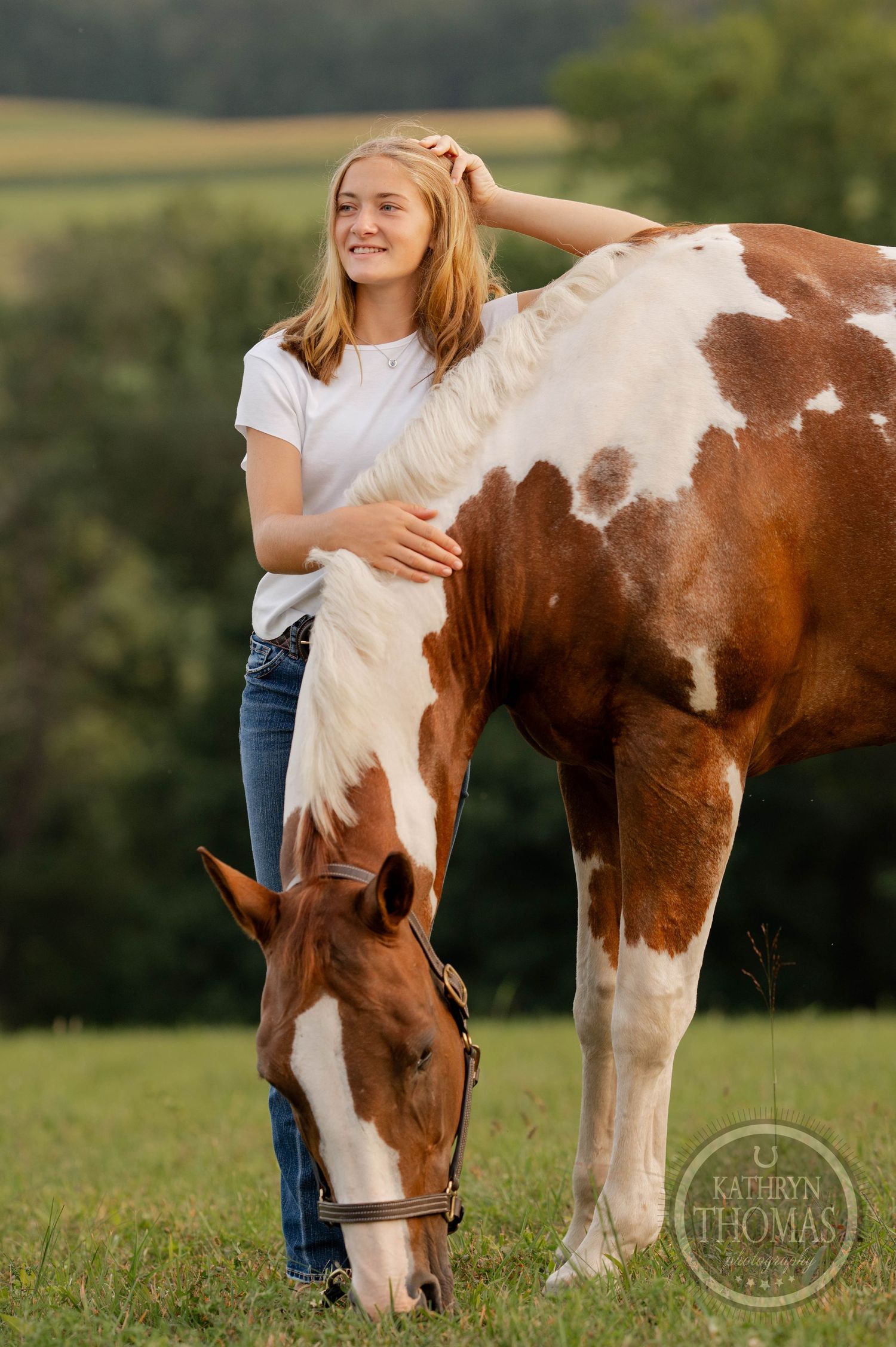 Person in white t-shirt embraces brown and white paint horse while standing in grassy field at sunset.