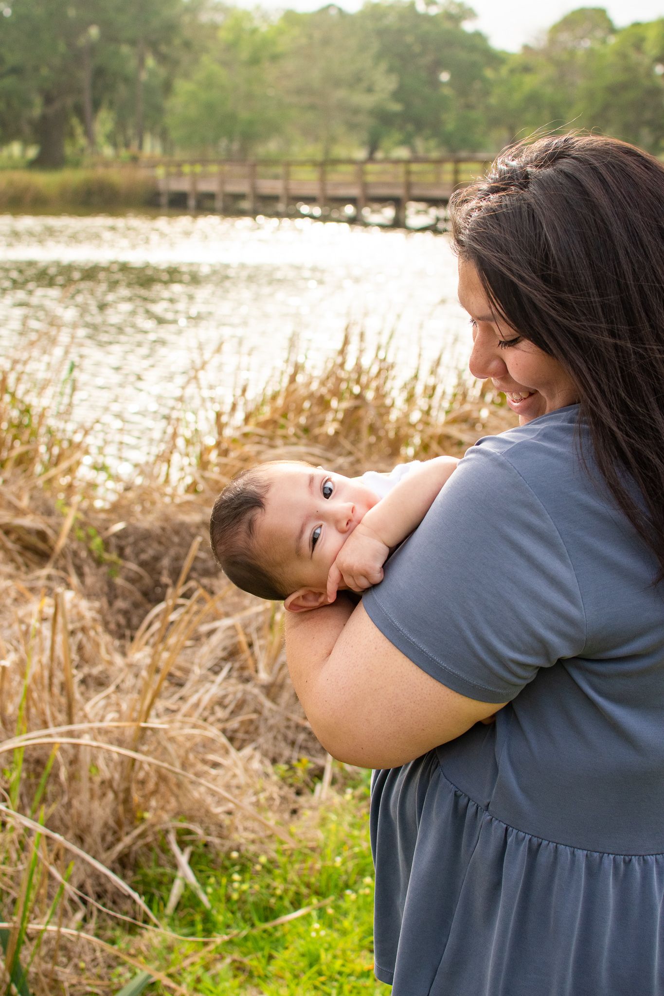 A tender moment captured by a lake with tall grass and a wooden bridge in the background.