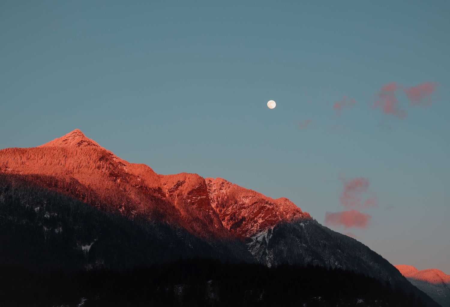 Full moon rises over snow-capped mountain peaks glowing orange in sunset light.