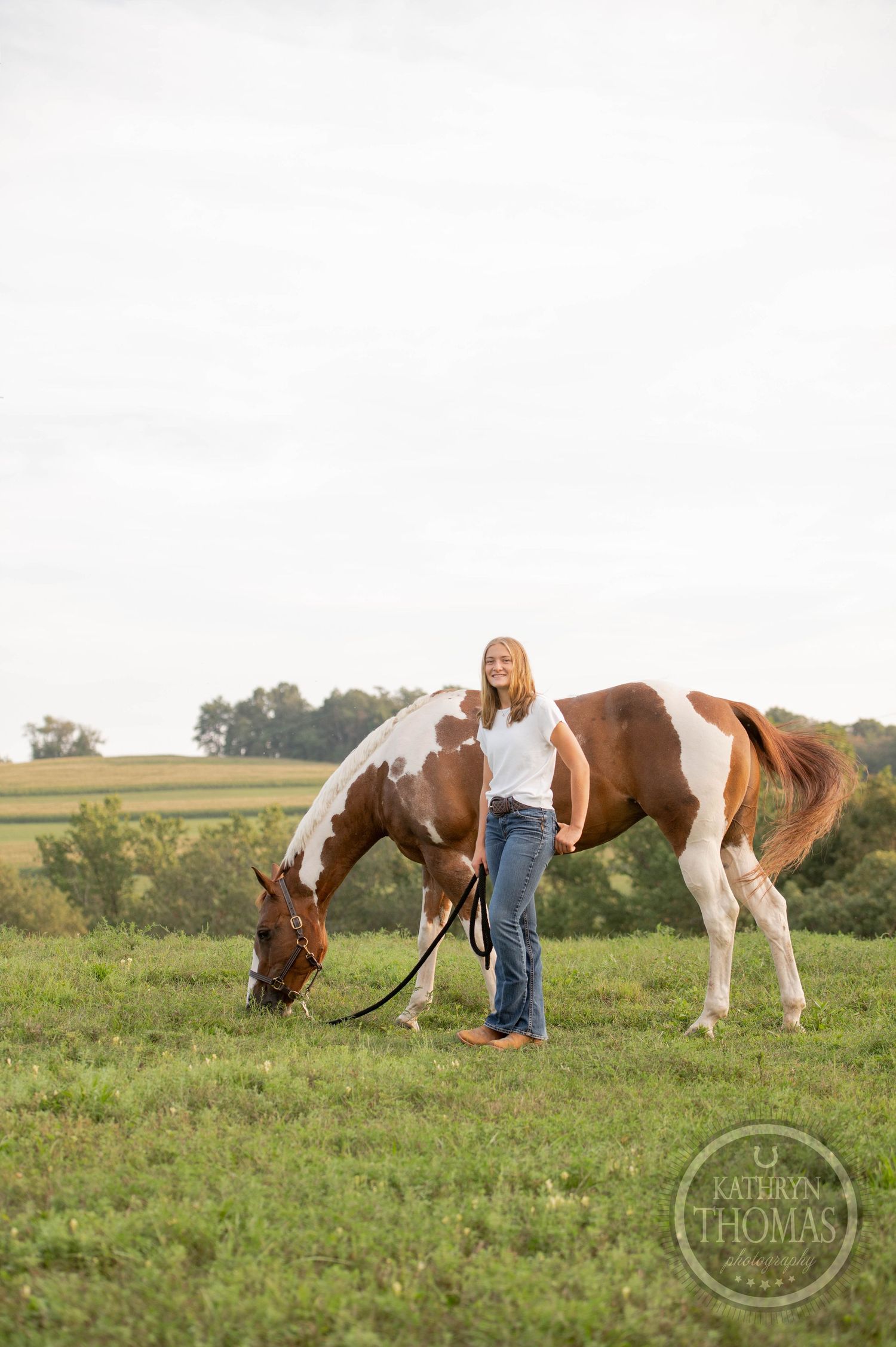 Paint horse grazing while person stands beside on grassy hilltop with pastoral view.