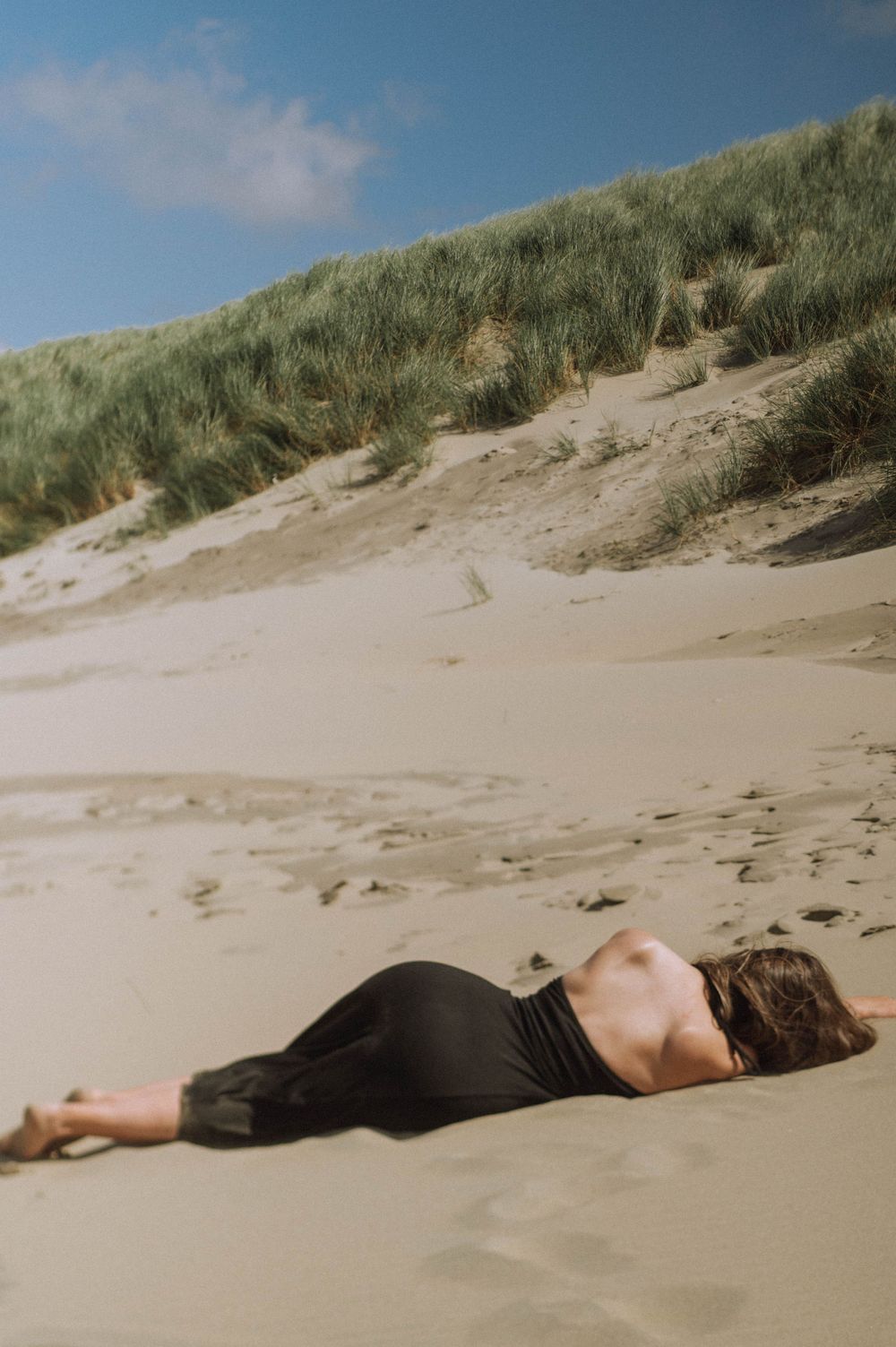 A figure in black clothing lies peacefully on sandy beach dunes with grass in the background.