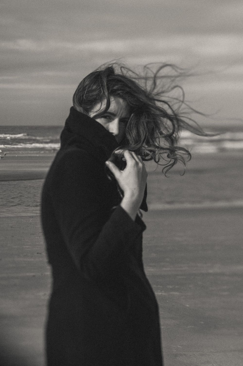 A figure in black stands against dramatic ocean waves in moody black and white photography.