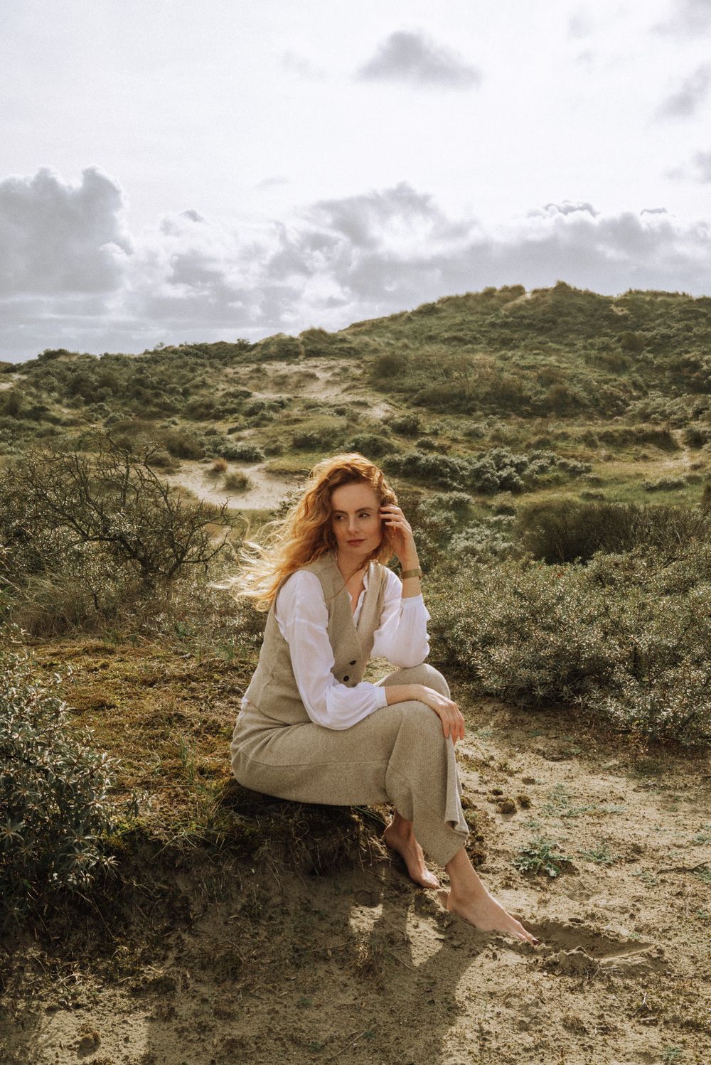 A person in light clothing sits contemplatively on sandy terrain surrounded by desert brush.