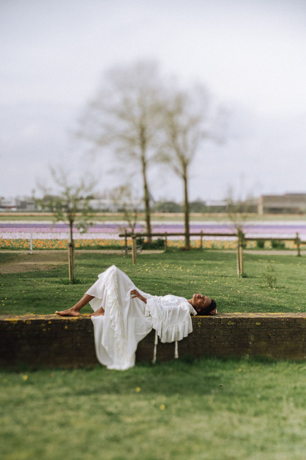 A figure in white lies artistically on grass with tulip fields in the distance.