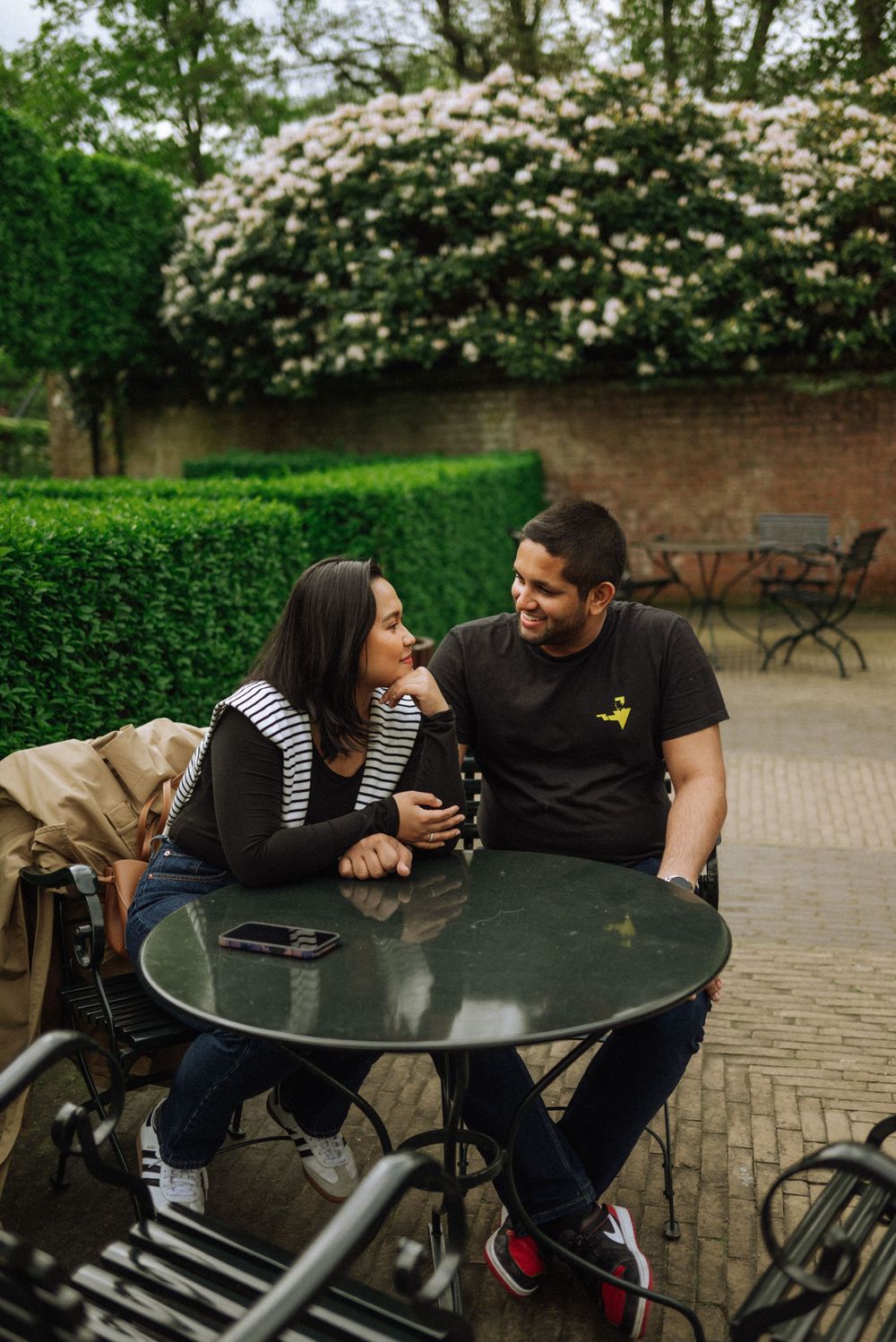 Two people enjoying conversation at a round table in a garden setting with blooming flowers.