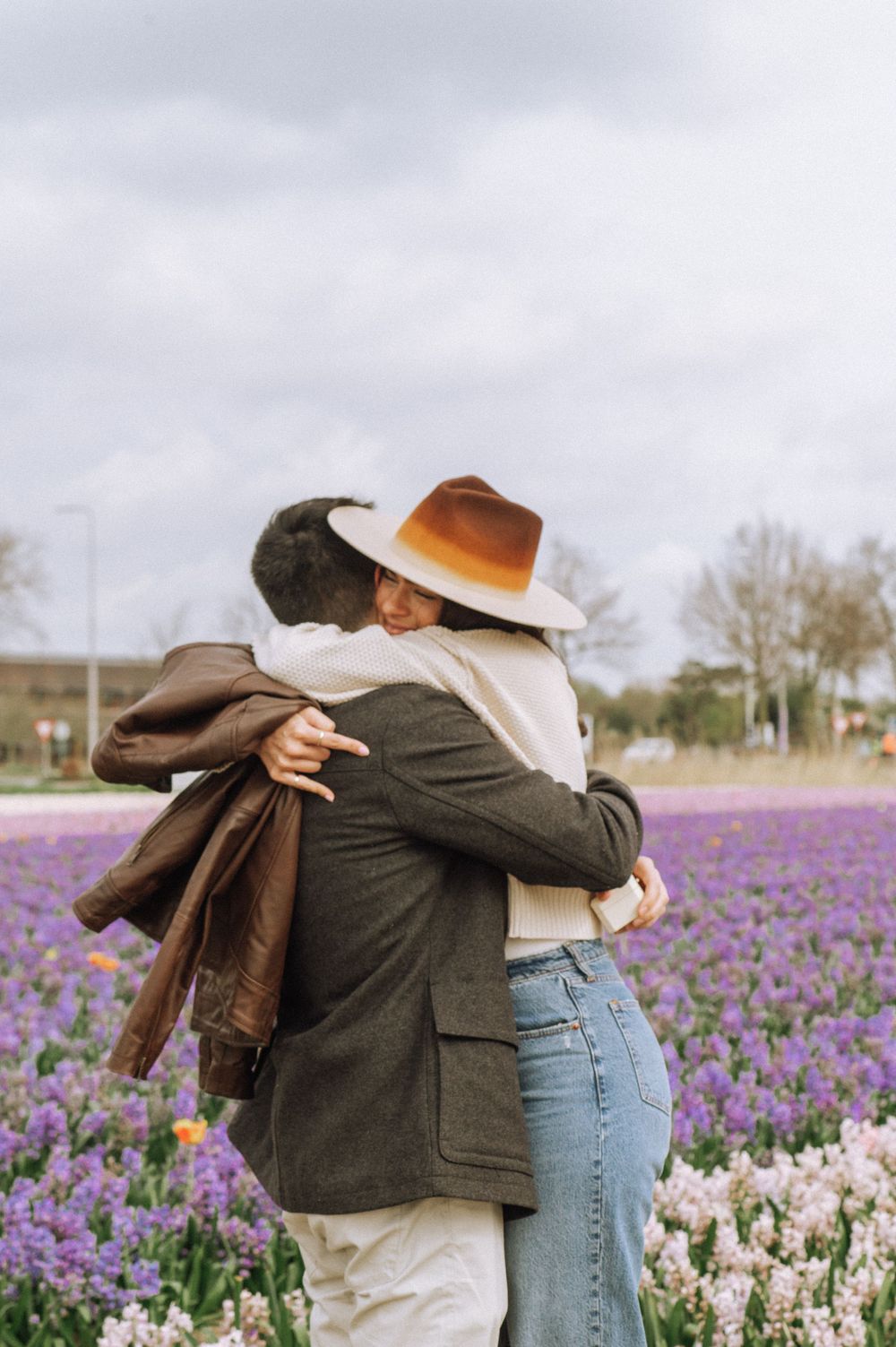 Two people embrace in a field of purple and white tulips under cloudy skies.
