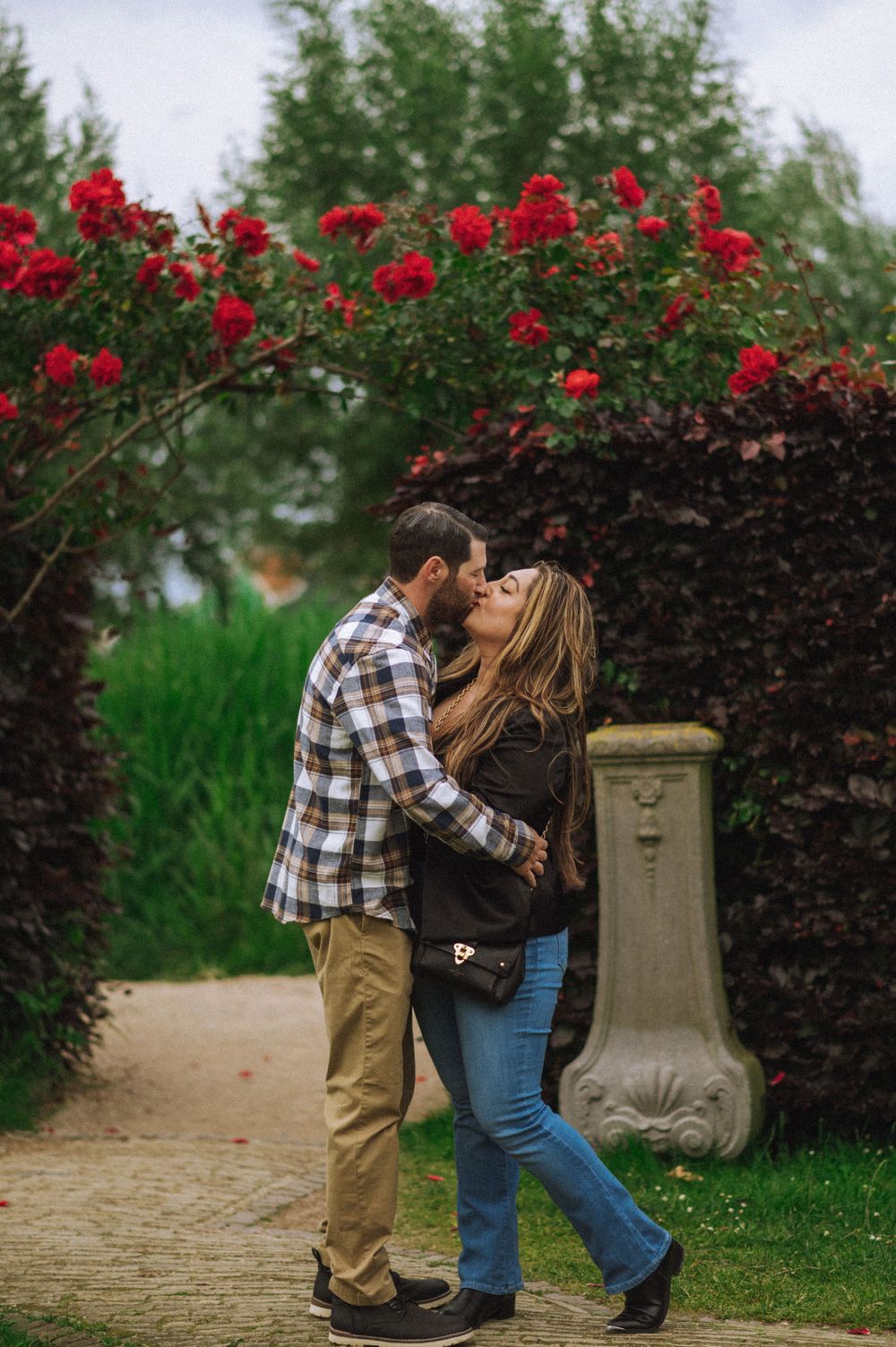 A couple embraces near red flowers and a decorative stone pillar in a garden.