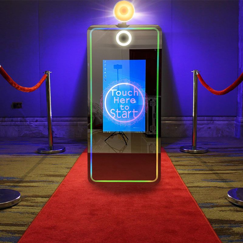 Photo booth setup with glowing screen and red carpet entrance ropes against blue lighting.
