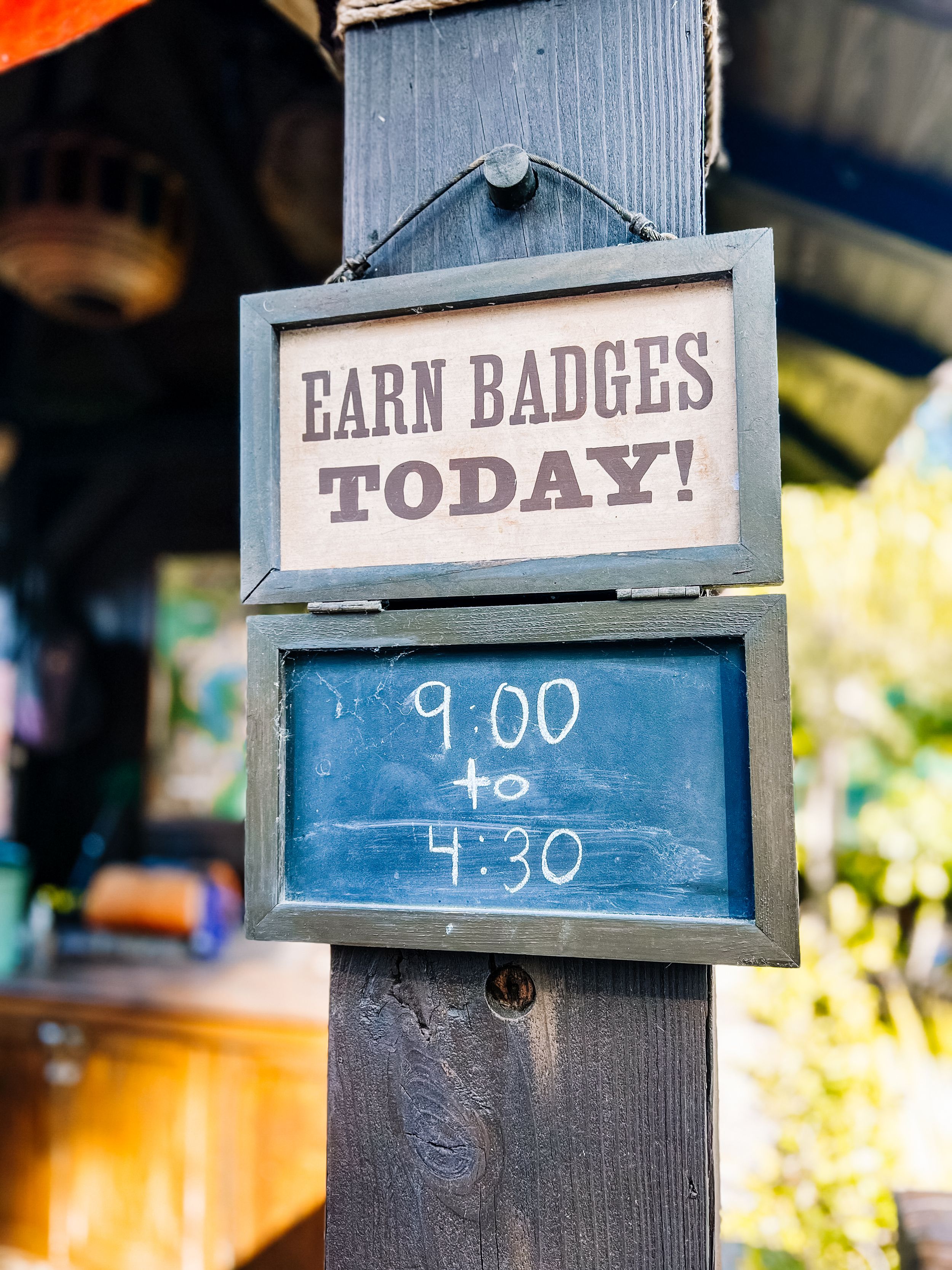 Wooden signs displaying operating hours and call to action text hang from posts with a blurred outdoor background.