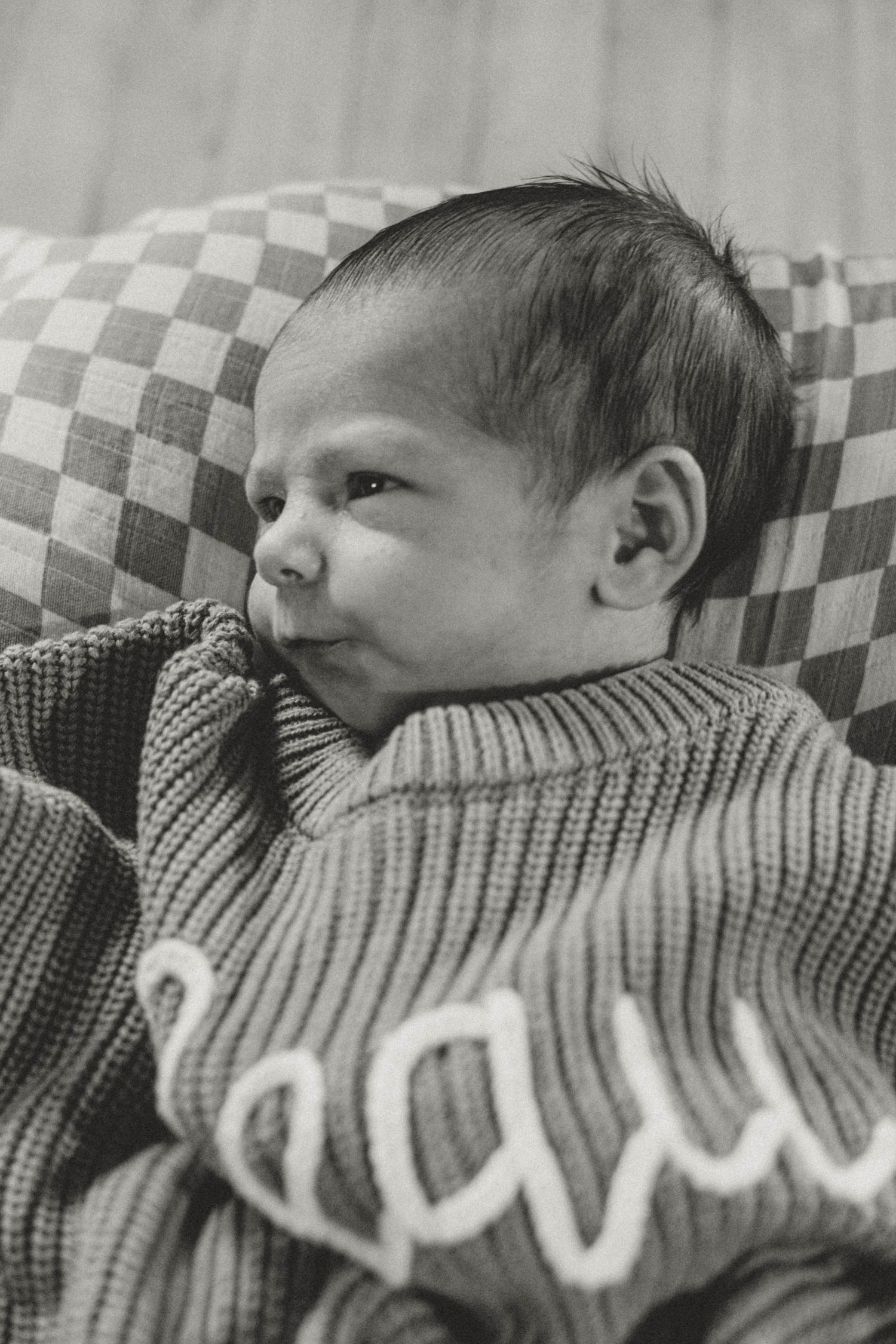 Black and white photo of a young baby wearing a knit sweater lying on a checkered cushion.