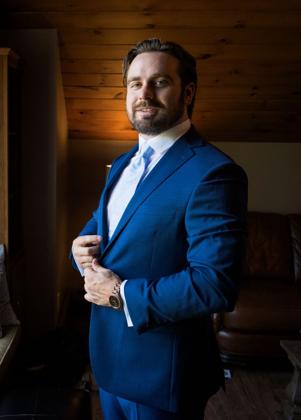Person in navy blue suit adjusting tie near window in rustic wooden room.