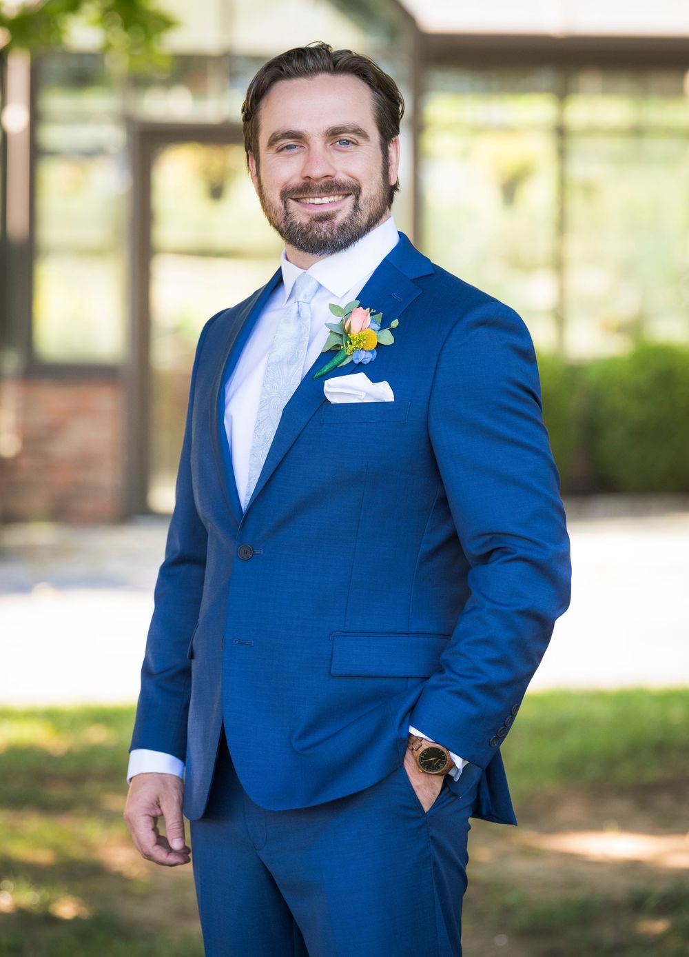 A groom wearing a royal blue suit and yellow boutonniere poses outdoors on his wedding day.