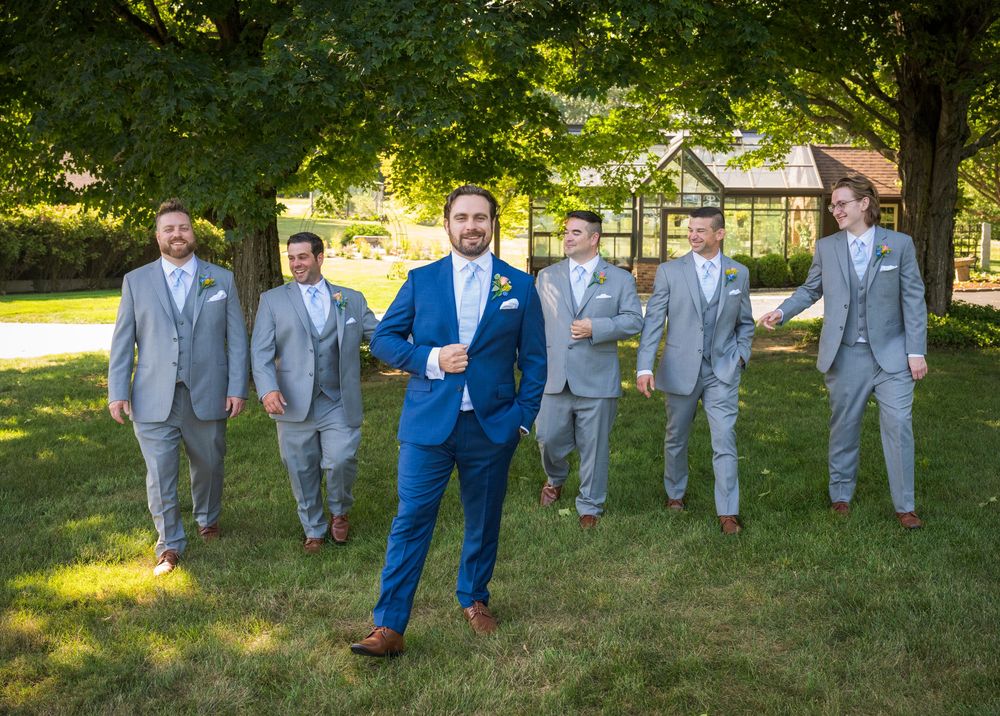 Groomsmen in gray suits and a groom in navy blue suit pose together on grassy lawn under trees.