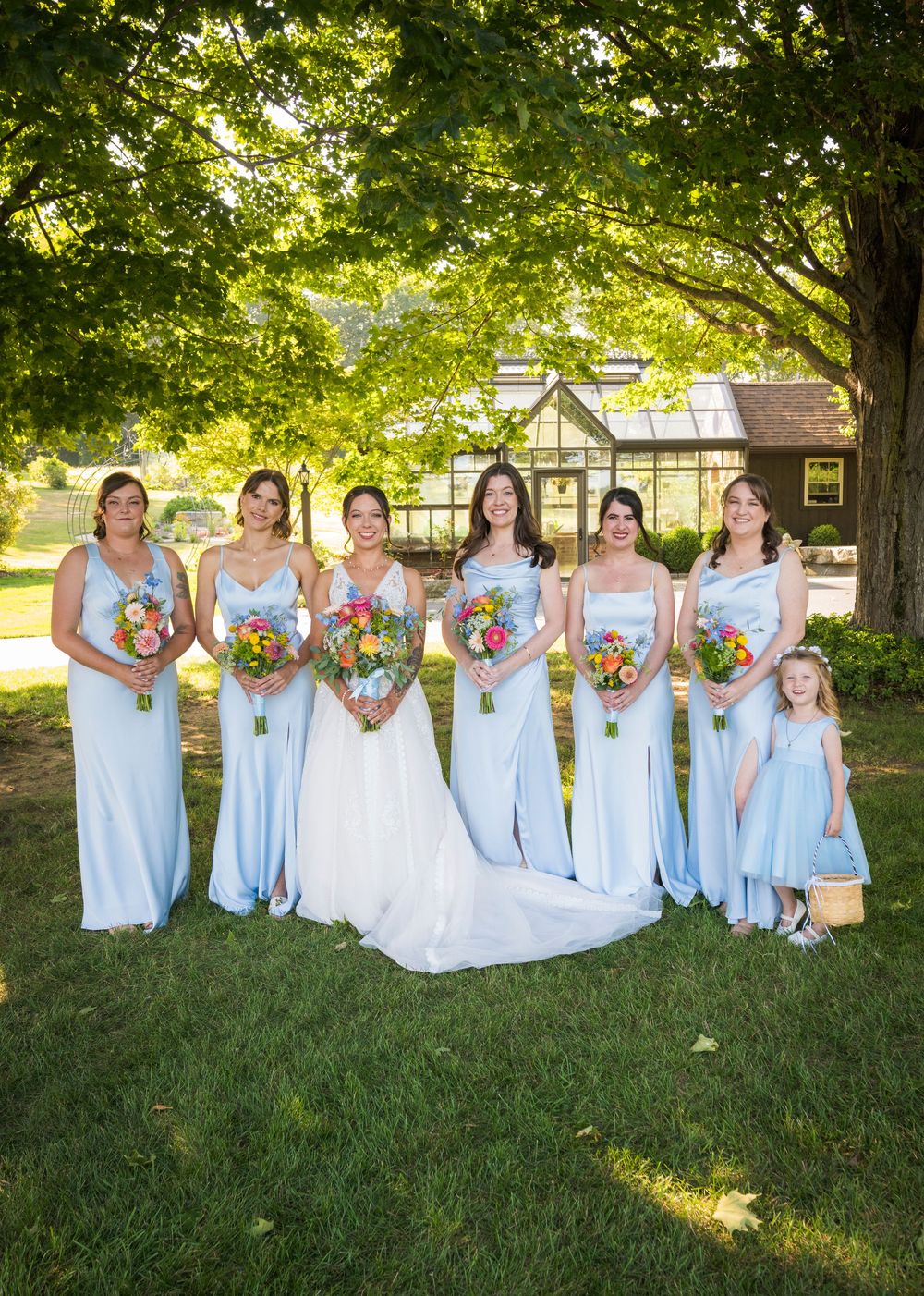 Bridesmaids in light blue dresses hold vibrant bouquets while posing on grass under trees.