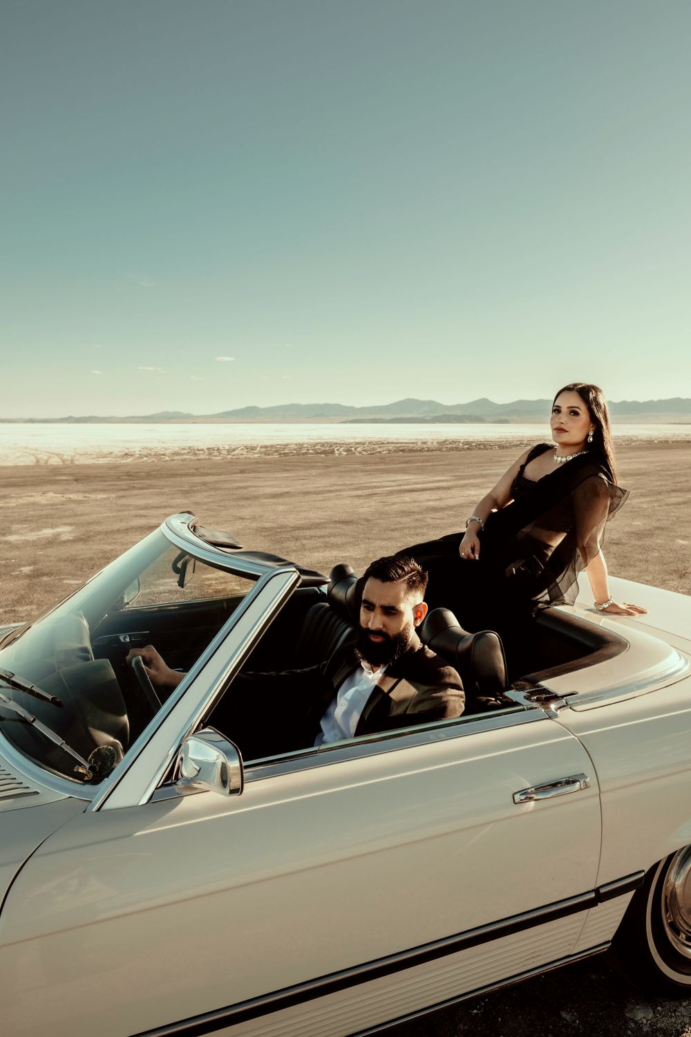 A vintage convertible car parked on a desert beach with mountains in the background at sunset.