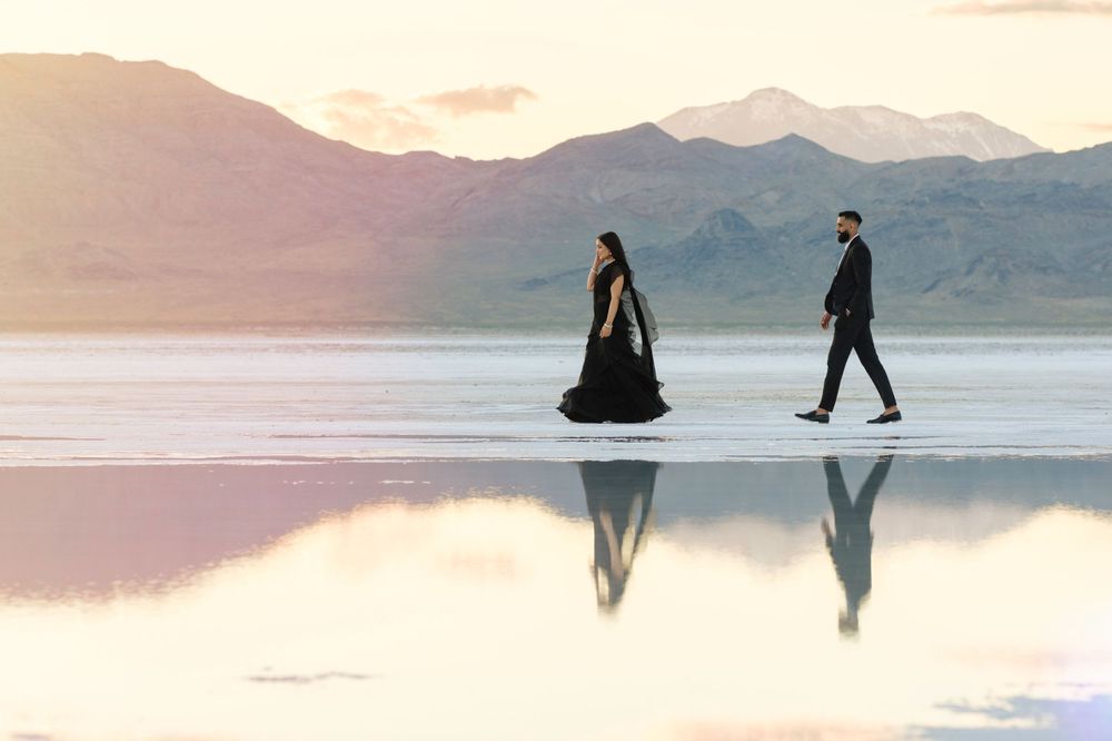 Two silhouettes walking on a reflective salt flat surface with mountains and sunset in the background.