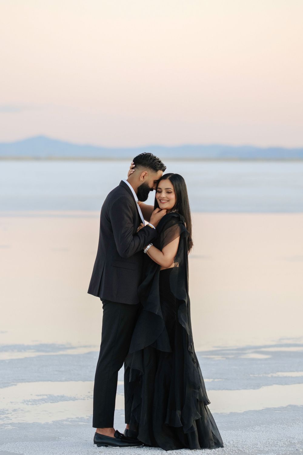 A romantic couple in formal black attire embraces on the white salt flats at sunset with mountains in the background.