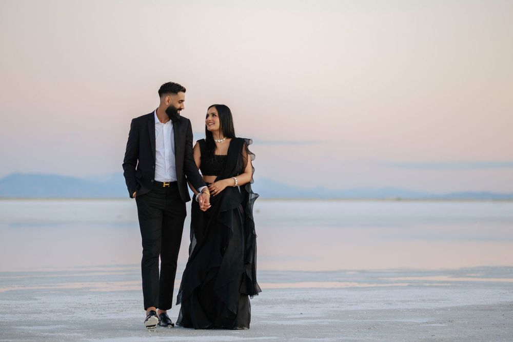 A couple in formal black attire walking together on salt flats with mountains in the distance.