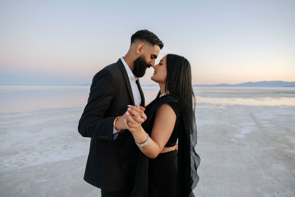 A couple shares a romantic moment while dancing on a salt flat at sunset, dressed elegantly in black attire.
