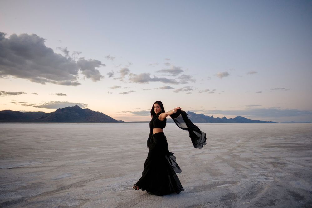 A person in a flowing black dress twirls on the Bonneville Salt Flats at sunset with mountains in the background.