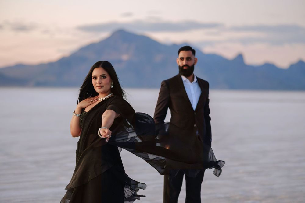 A couple in elegant black outfits posing on salt flats with dramatic mountain backdrop at dusk.
