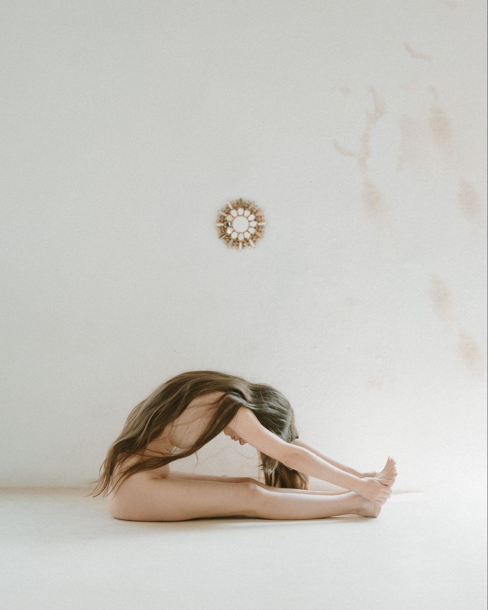 Person practicing yoga in seated forward fold pose against white wall with a decorative sunburst mirror.