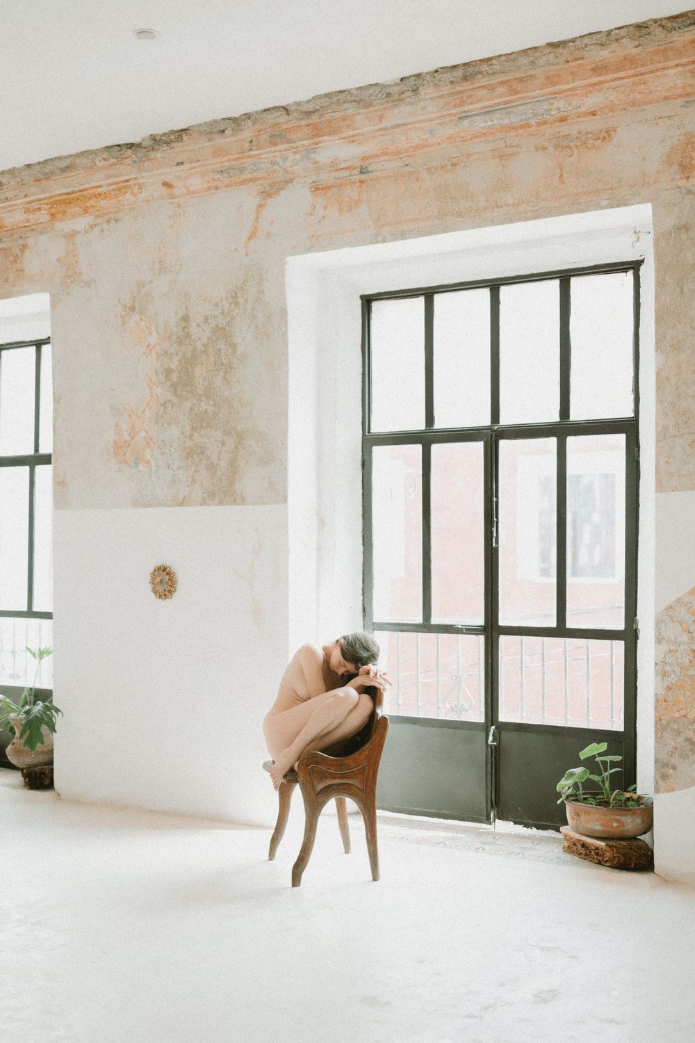 Person in brown coat sitting on minimalist chair by large window in white room with distressed walls.