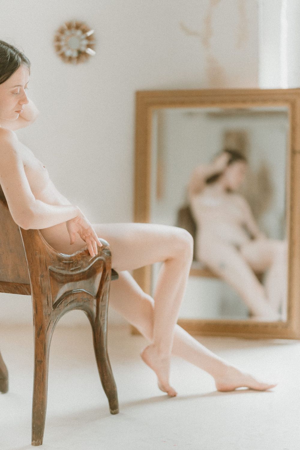 Soft, artistic boudoir photography features someone seated on an antique wooden chair with a mirror reflection in background.