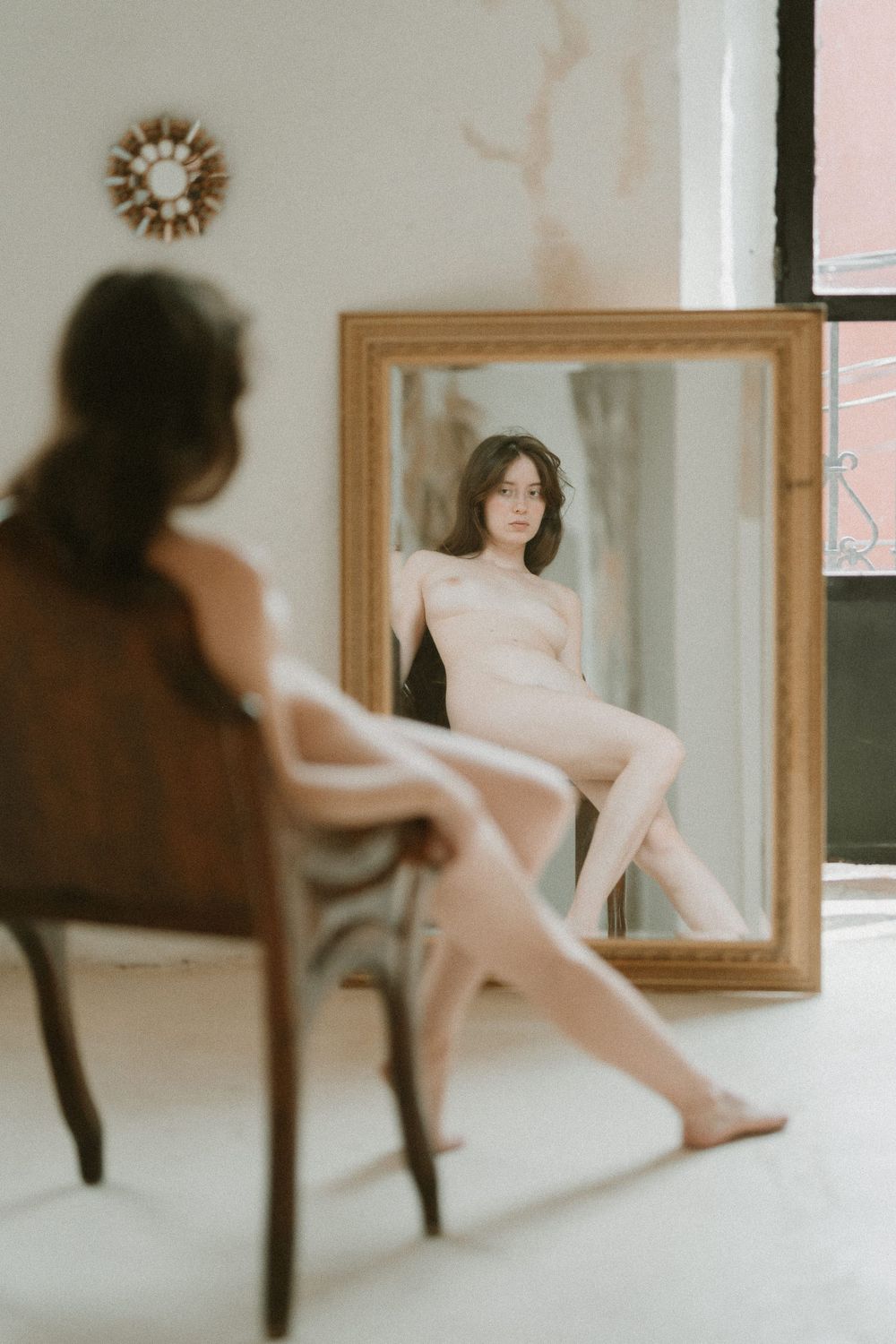 A soft, dreamy reflection in a vintage gold-framed mirror creates an artistic boudoir scene in muted tones.