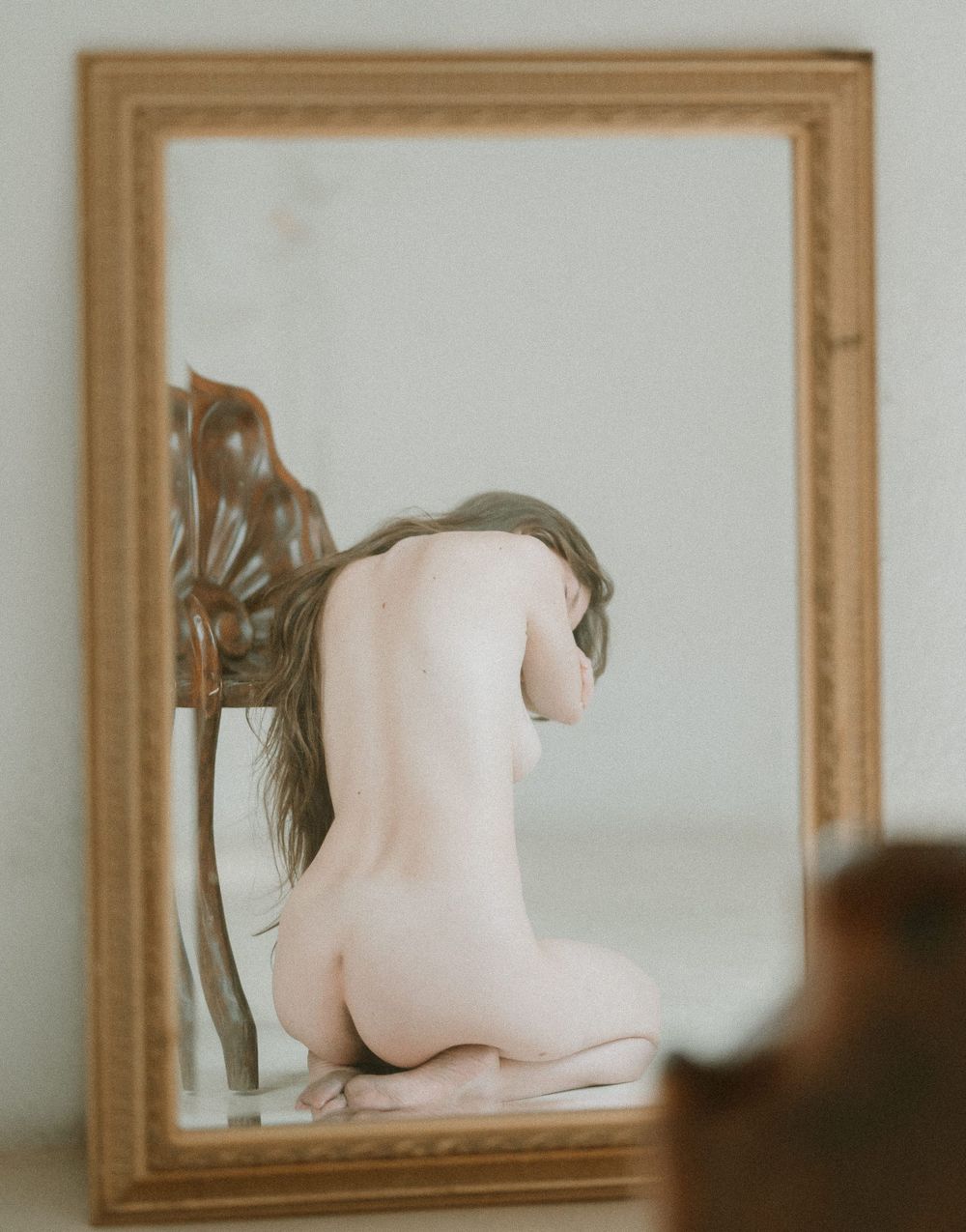 Artistic nude figure reflected in a vintage wooden framed mirror against a neutral background.
