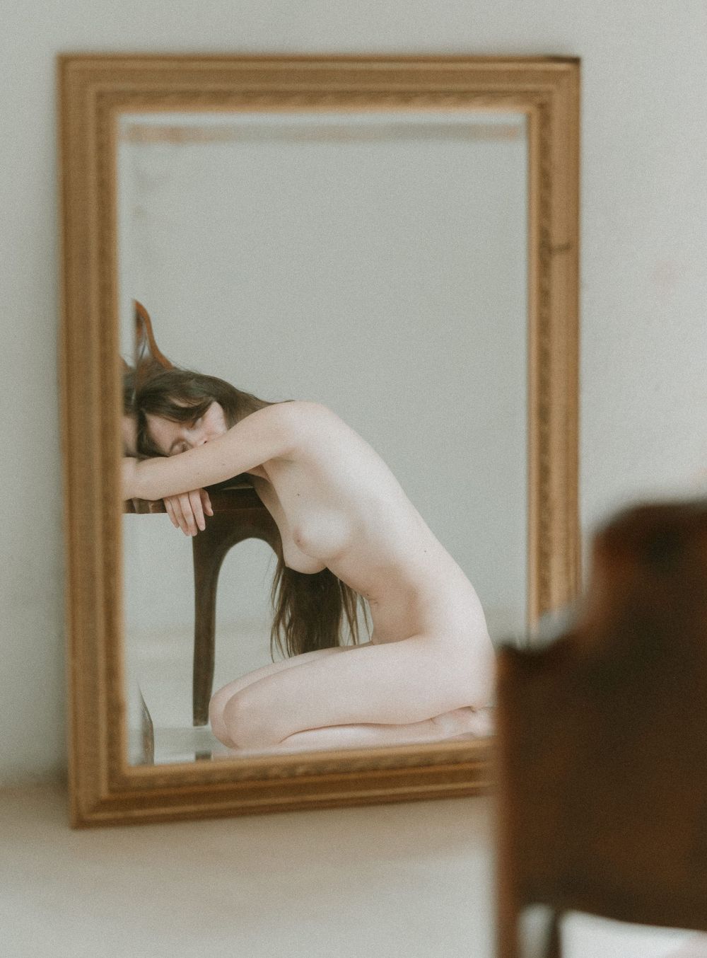 Artistic nude photograph showing a silhouette reflecting in a gold-framed mirror against a light colored wall.