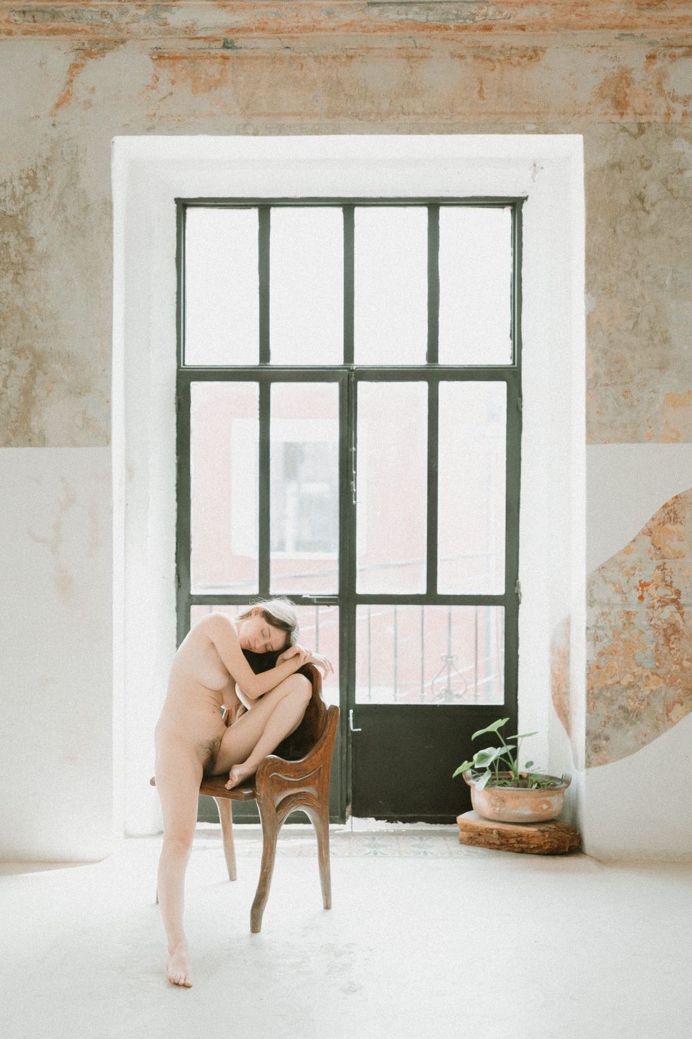 Minimalist scene with a wooden chair by a large industrial window and potted plant in a bright white room.