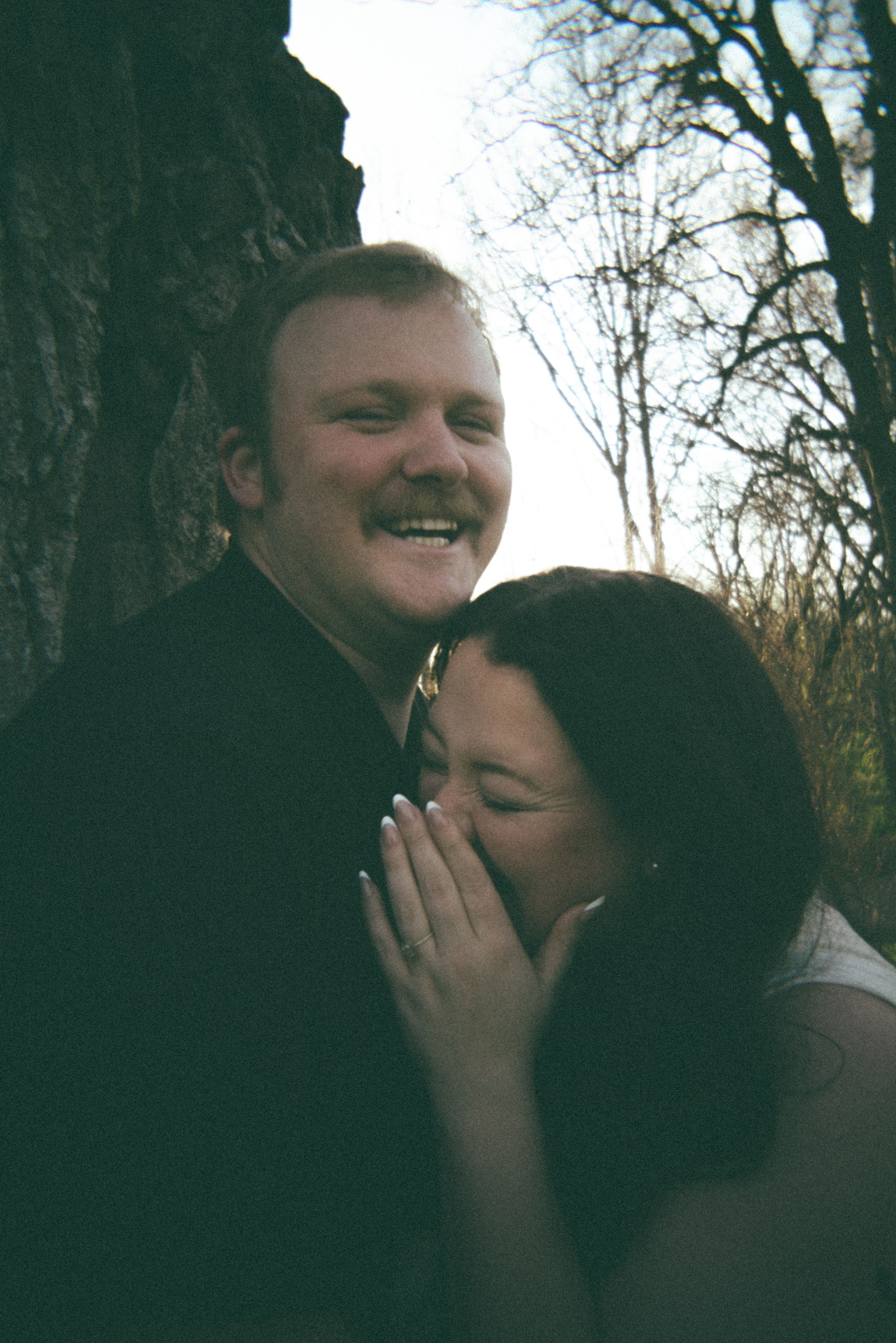 A laughing couple shares a tender moment leaning against a tree outdoors in a romantic engagement photo.