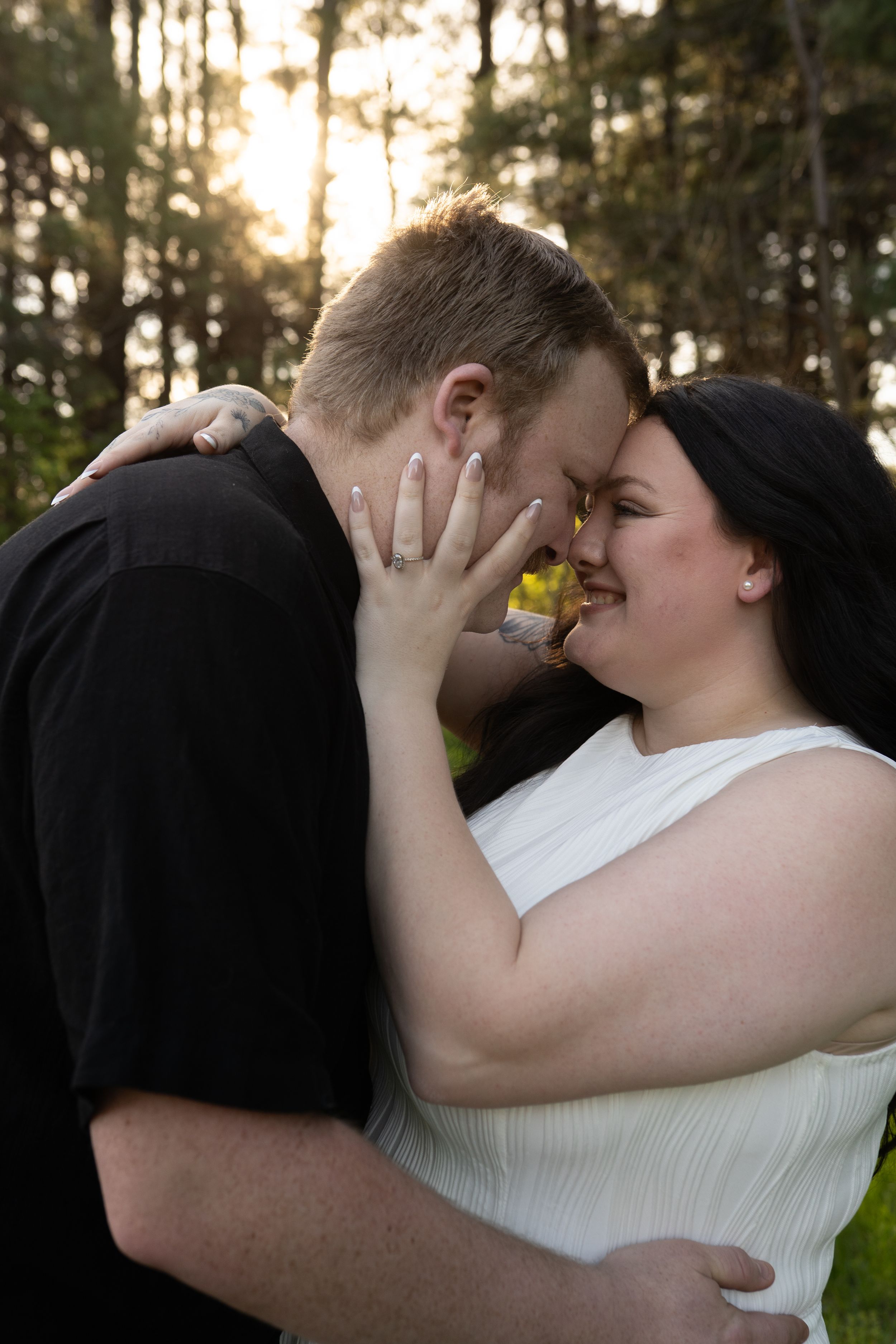 Couple sharing a romantic forehead touch outdoors, woman in white dress showing engagement ring, surrounded by trees.