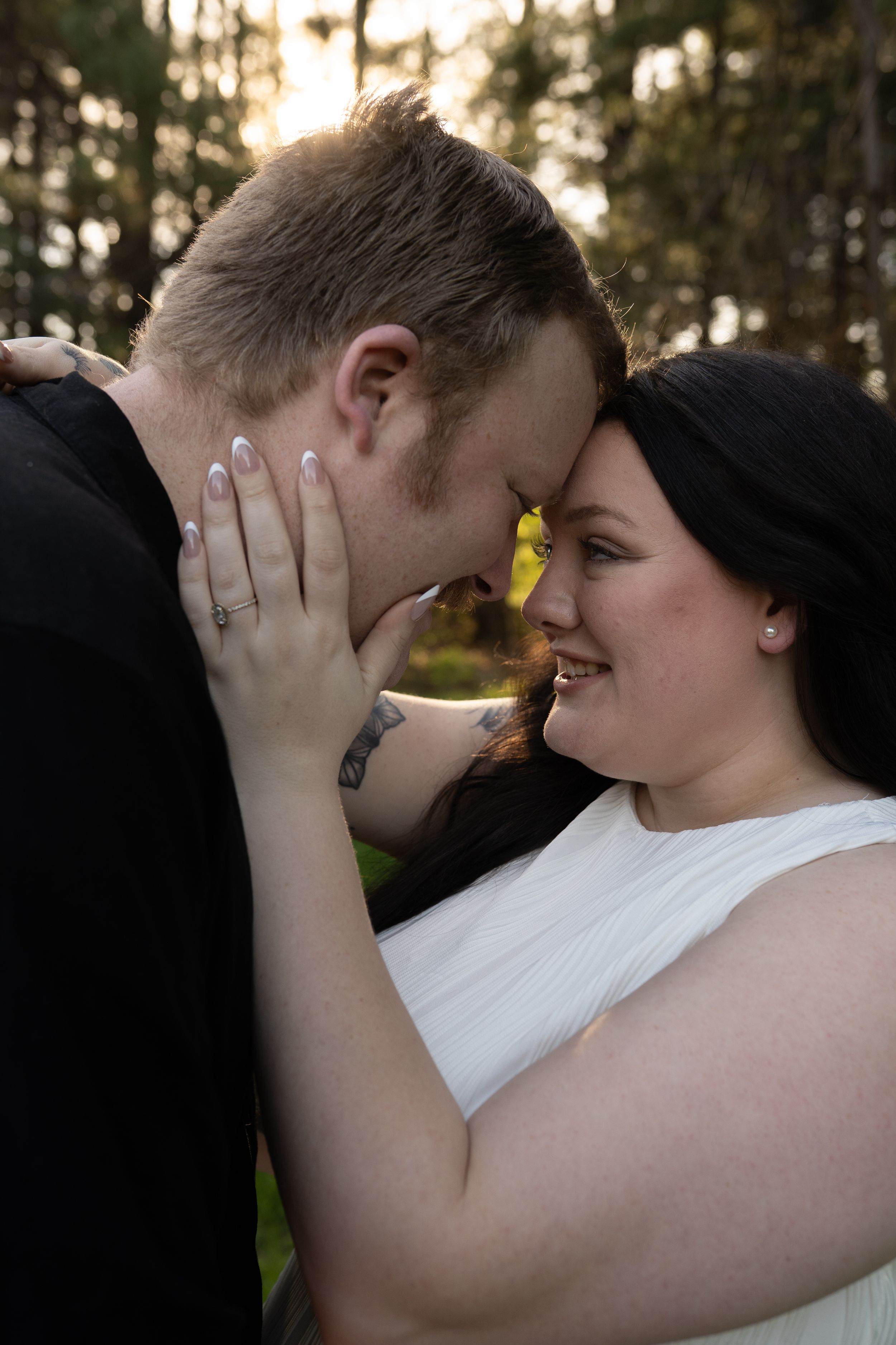 Smiling bride in white dress touches groom's face as they lean foreheads together outdoors at golden hour.