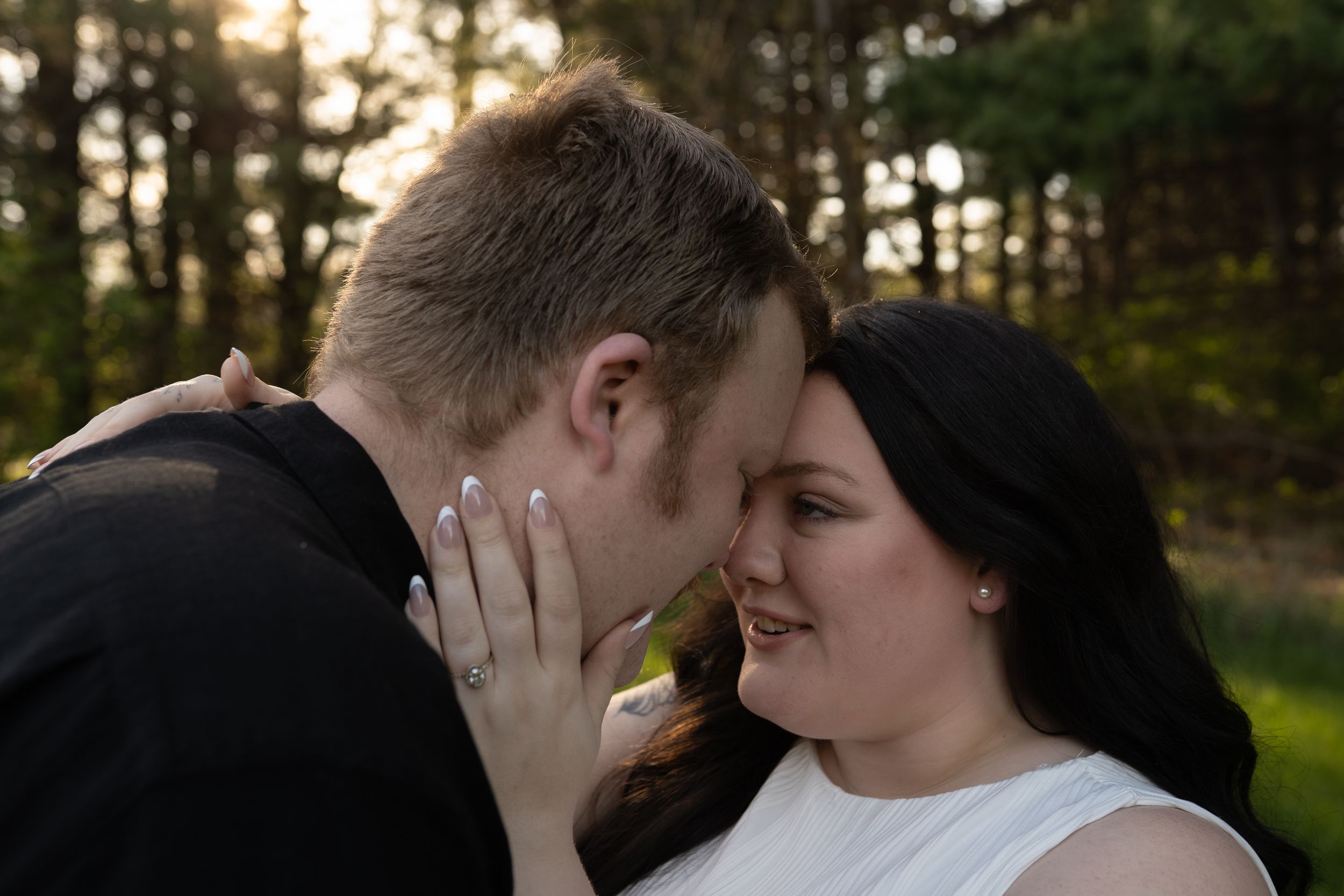 Couple sharing a romantic moment outdoors, woman smiling with engagement ring visible, forest backdrop at golden hour.