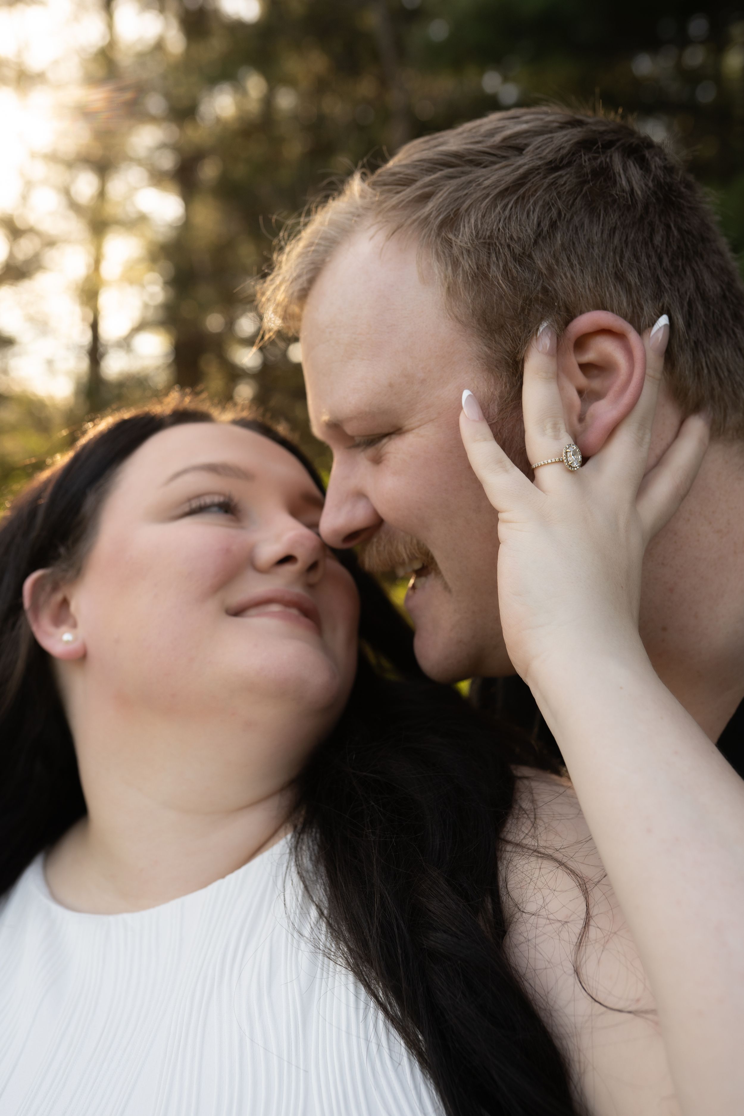 A couple shares a tender nose-to-nose moment outdoors, woman with dark hair wearing engagement ring gently holds mans face.