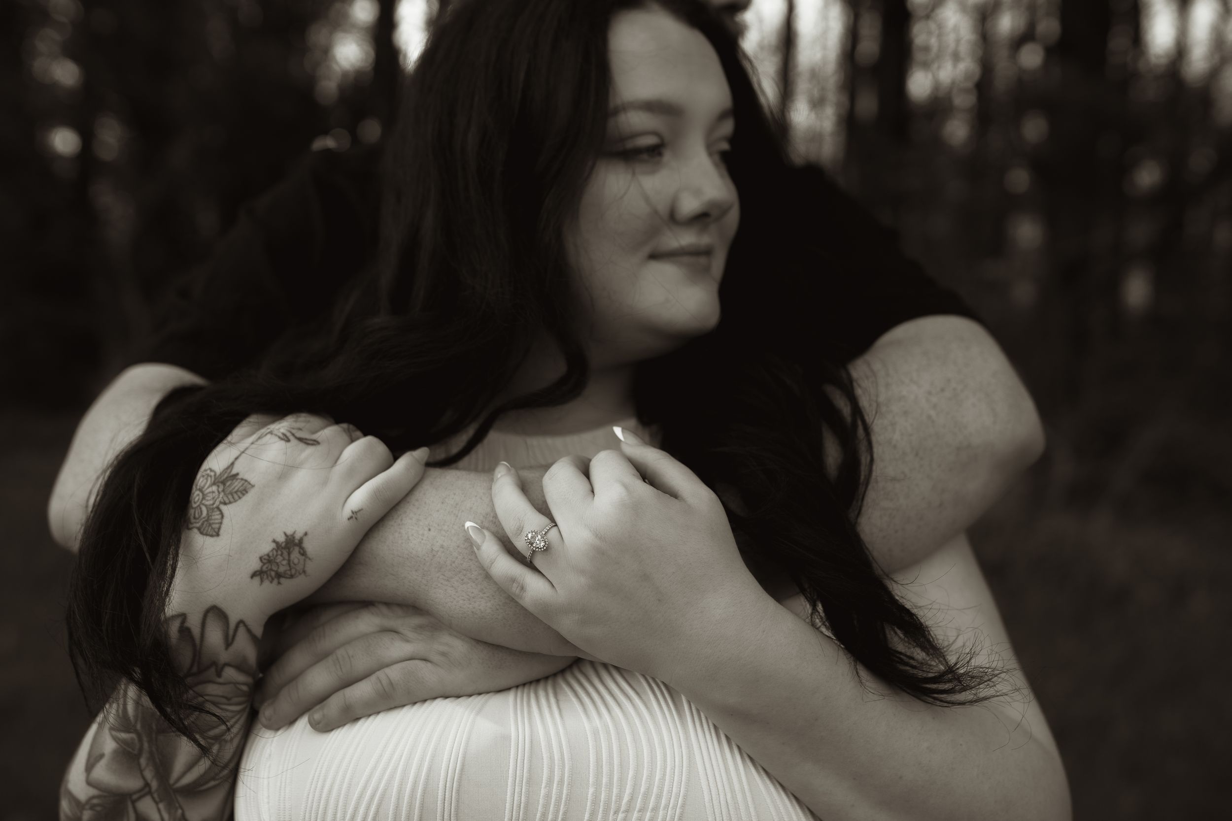 Black and white photo of two women embracing tenderly outdoors, one with tattoos and a ring visible.