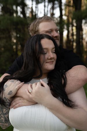 A man embraces a woman with dark hair and tattoos outdoors in a forest setting during an engagement photo session.